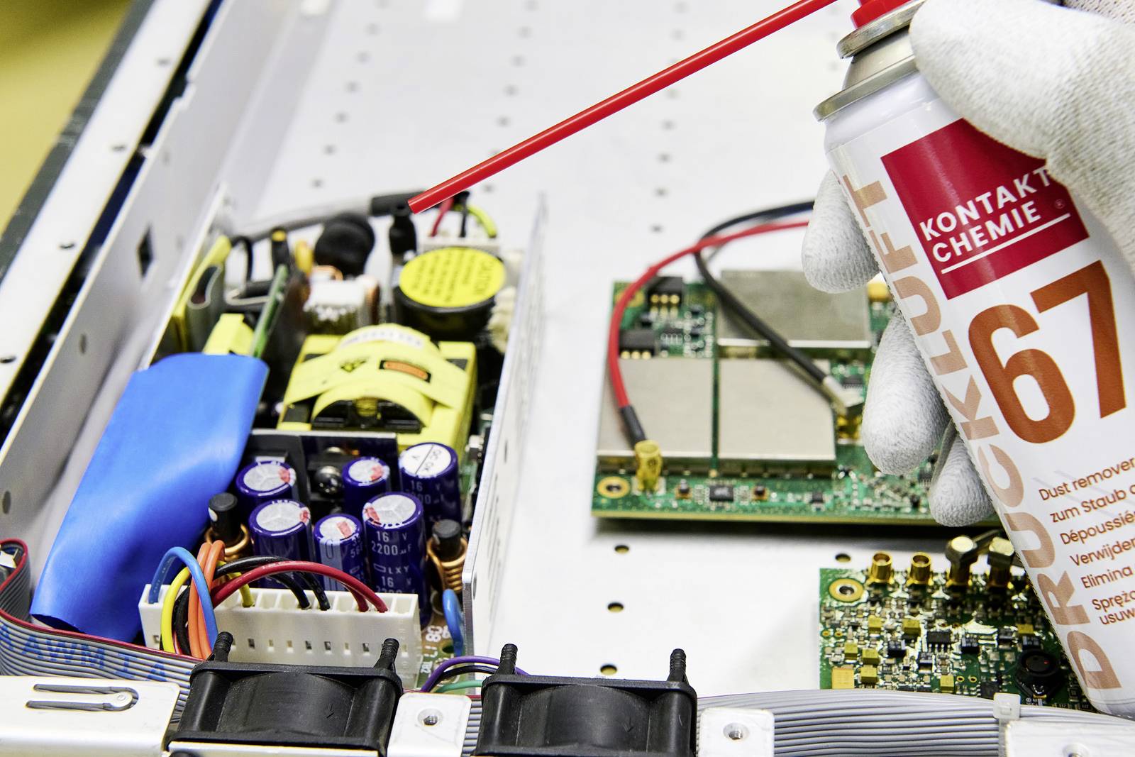 Osoba čistí vnitřek elektronického zařízení pomocí spreje. Jsou viditelné kondenzátory a elektrické obvody.