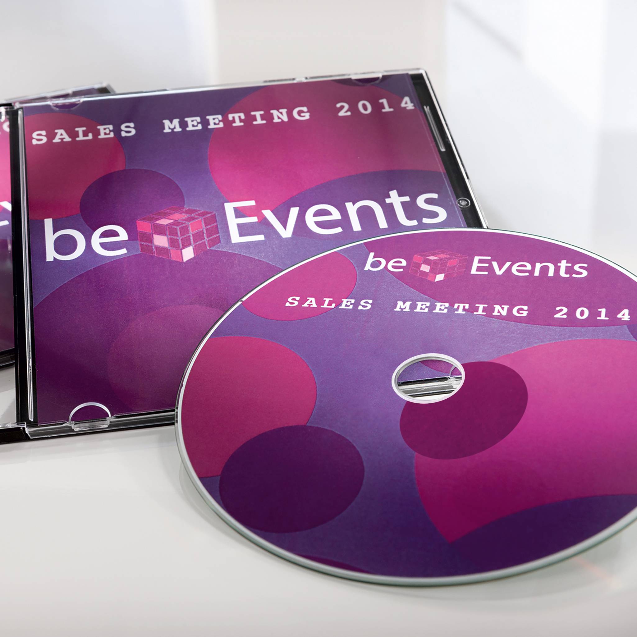 CD disky s nápisem 'Sales Meeting 2014' a 'beEvents', ozdobené růžovou grafikou. Leží na stole.