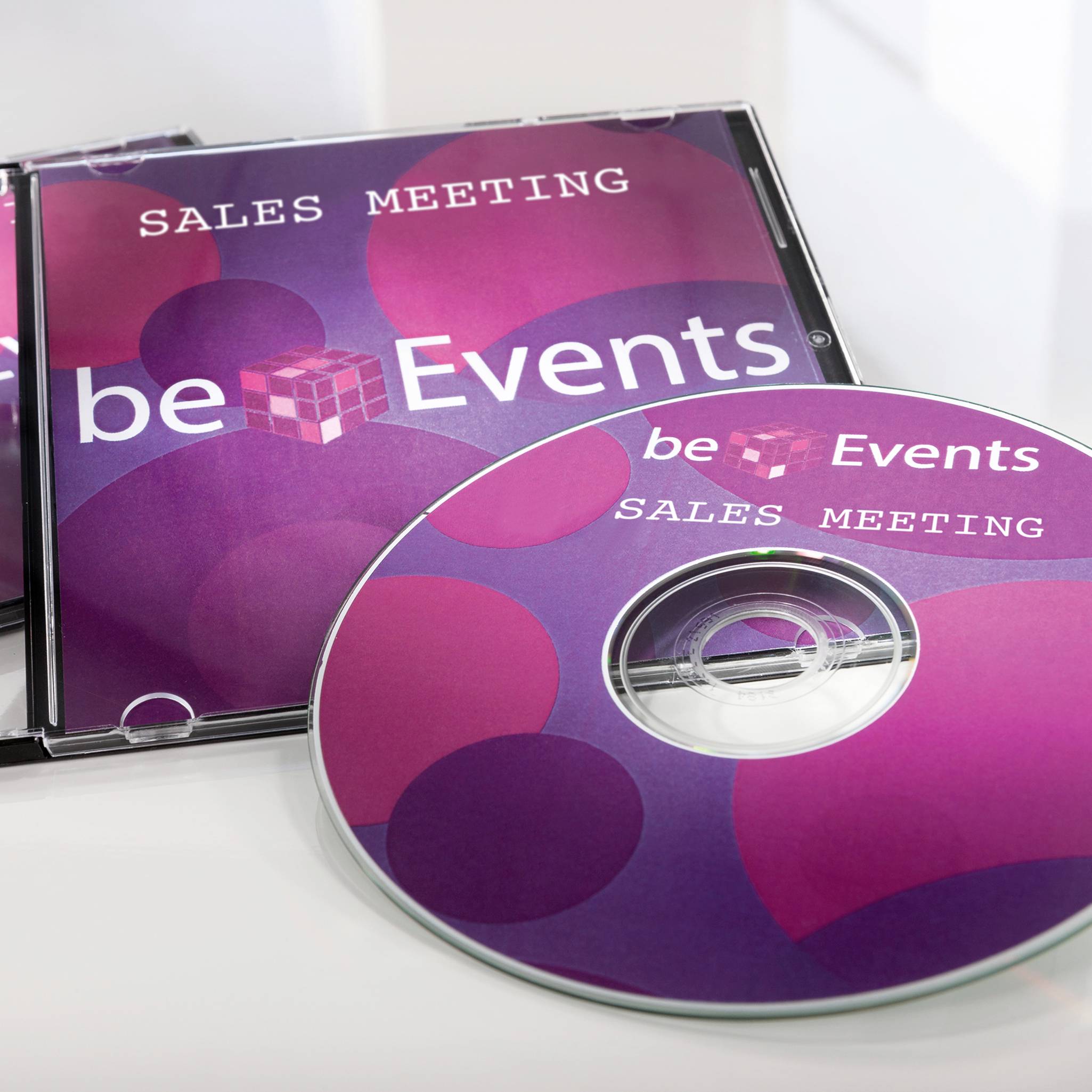 CD obaly s nápisem 'Sales Meeting Be Events' na růžovo-fialovém pozadí s kruhovými a krychlemi vzory.