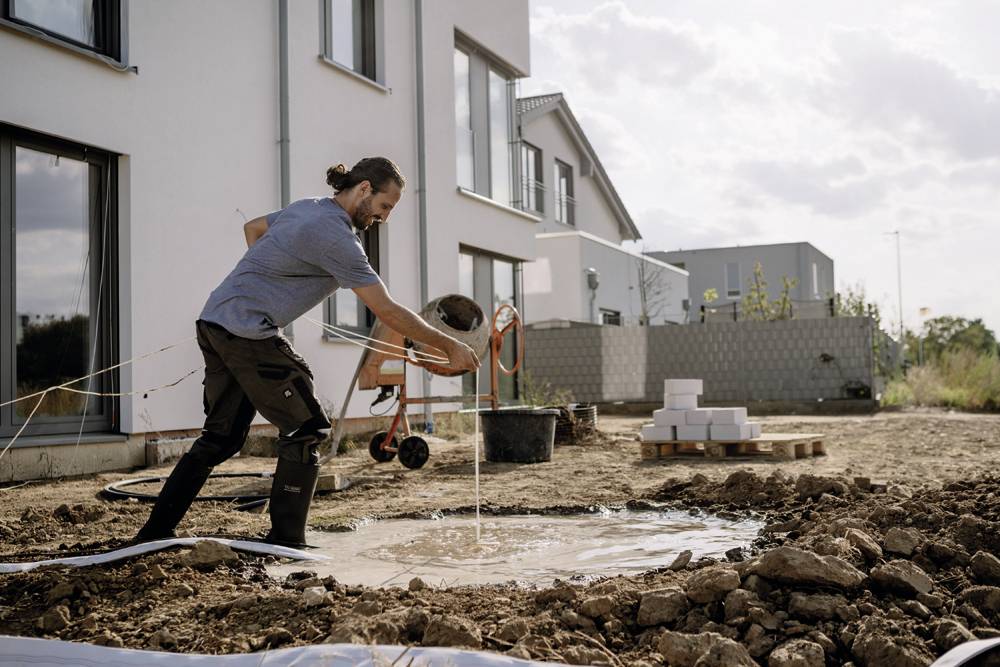 Muž zalévá vodu na půdu částečně zastavěné plochy vedle moderního domu. V pozadí jsou vidět další budovy.