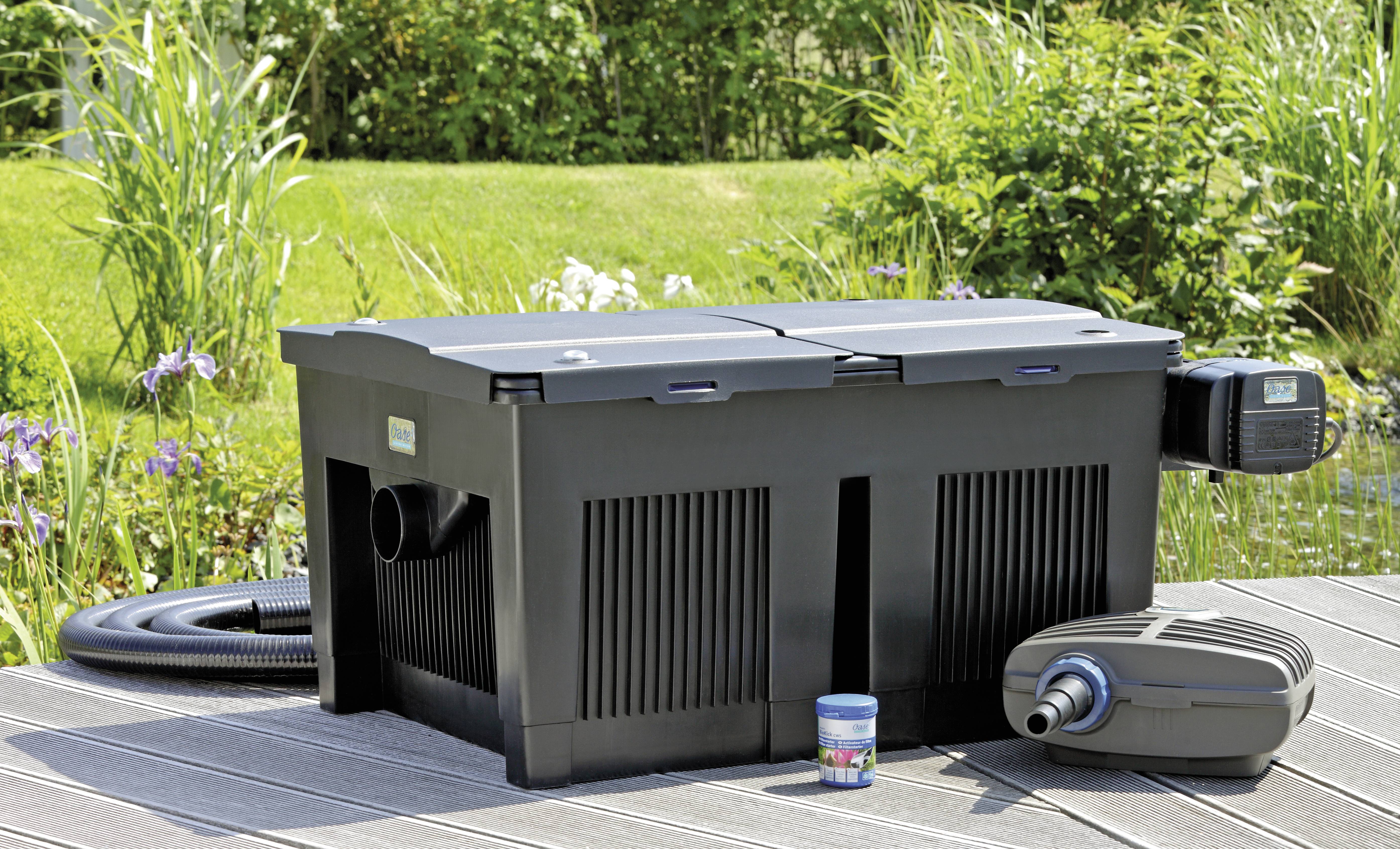 Černý filtrační systém pro rybníček s hadicemi a příslušenstvím je umístěn na dřevěné terase s rostlinstvím v pozadí.