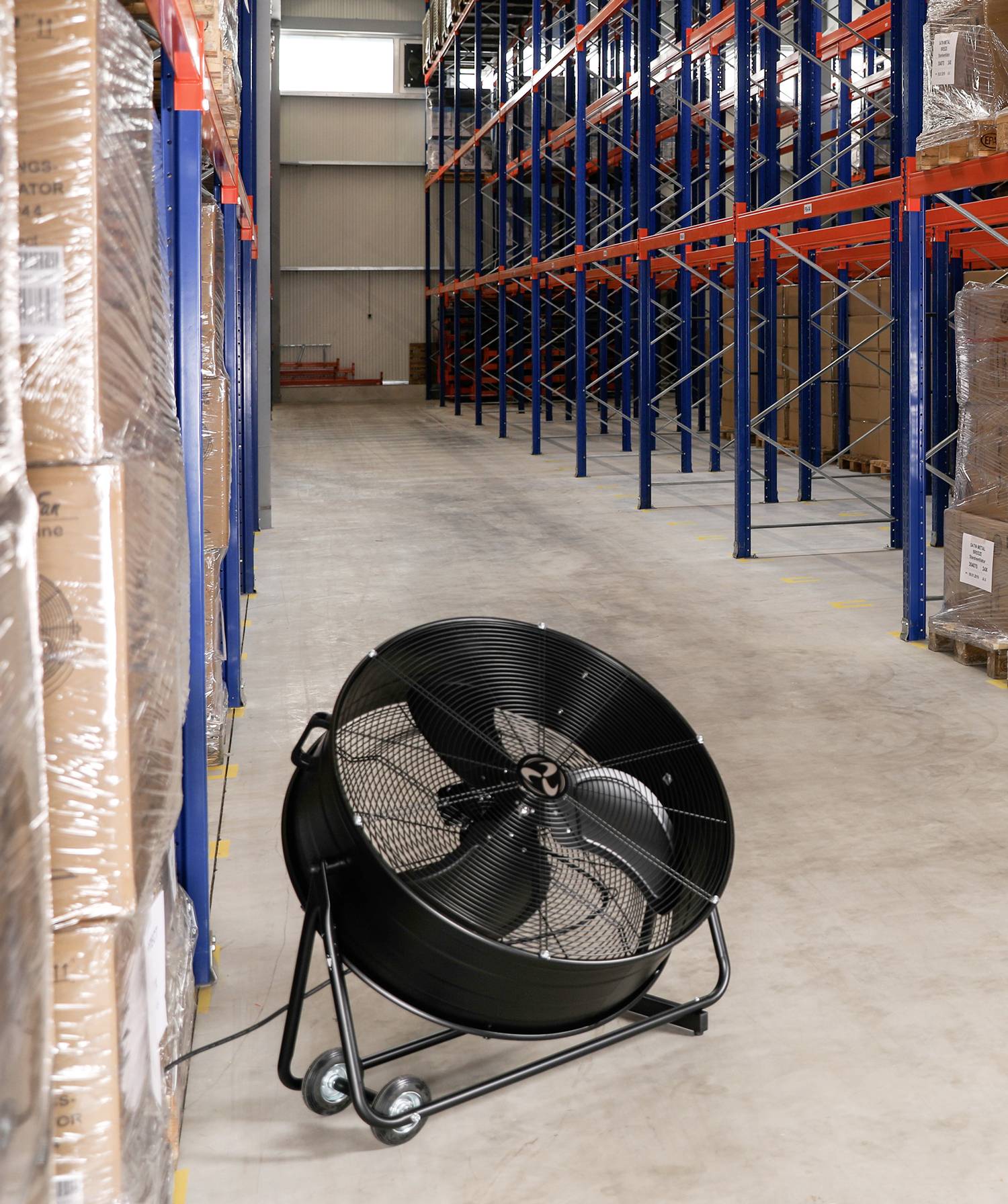 Ventilátor ve skladu, umístěný před stohy složených krabic a prázdnými regály. Optimalizuje cirkulaci vzduchu.