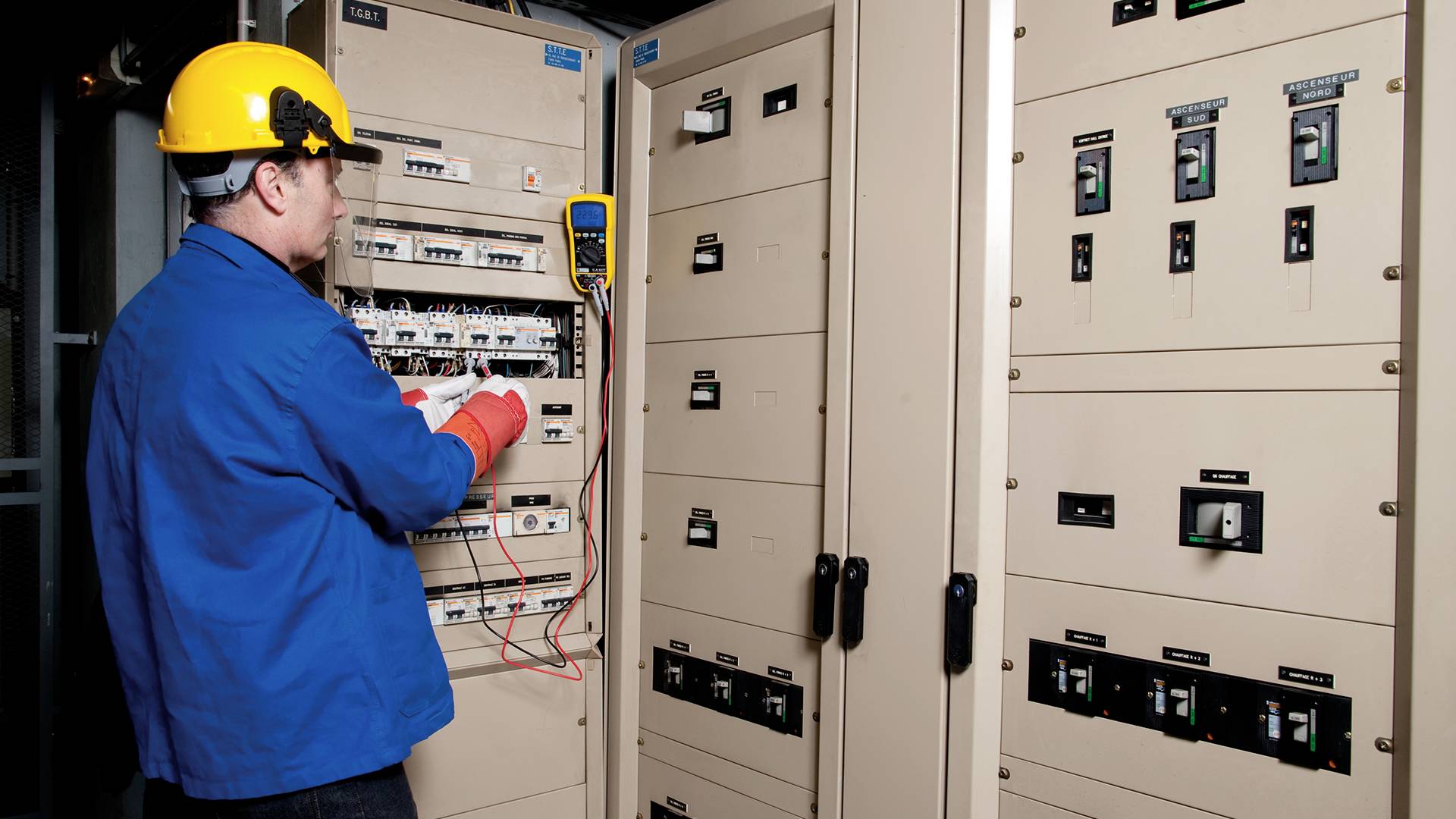 Elektrikář v modré pracovní kombinéze a žluté přilbě kontroluje rozvaděče multimetrem v řídicí místnosti.