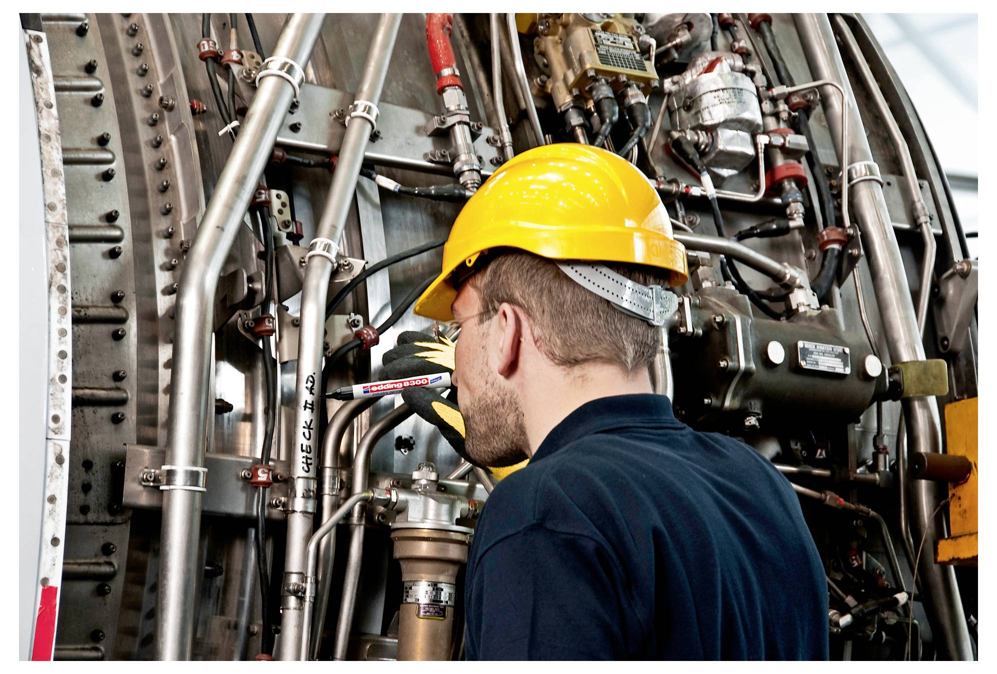 Pracovník v žluté přilbě kontroluje husté průmyslové potrubí a zařízení uvnitř továrny.