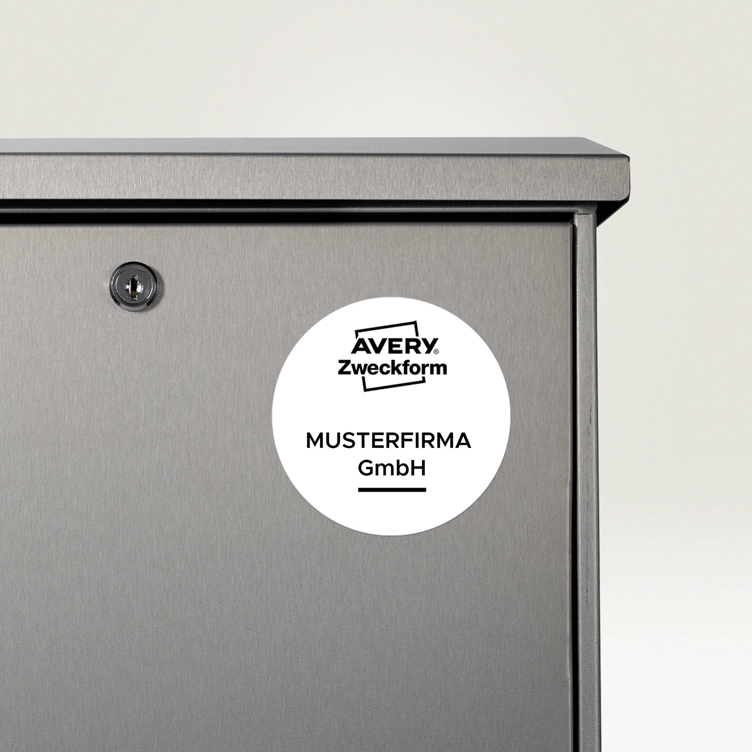 Šedý kovový poštovní schránka s kulatou nálepkou na přední straně. Na nálepce je napsáno 'Avery Zweckform' a 'Musterfirma GmbH'.