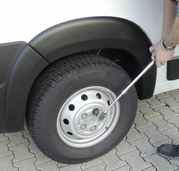 Osoba povolující kolové matice na pneumatice automobilu pomocí klíče, demonstrující postup výměny kola. Vozidlo je zaparkované na dlažebních kostkách.