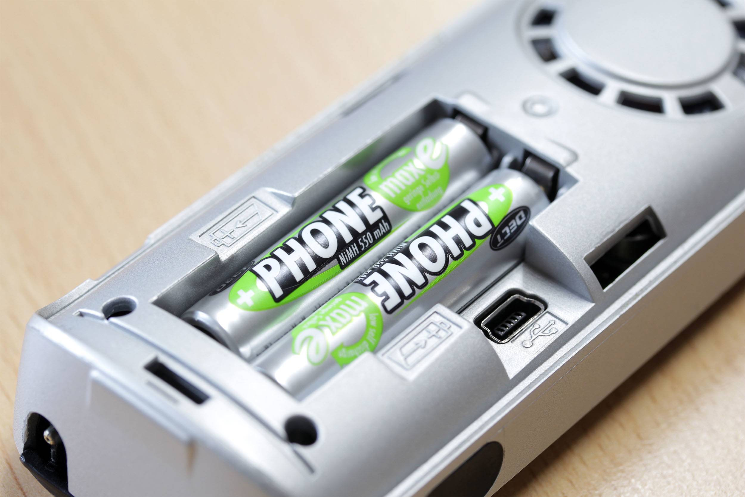 Stříbrné elektronické zařízení s otevřeným prostorem pro baterie, do kterého jsou vloženy dvě zelené baterie AA s nápisem "PHONE".