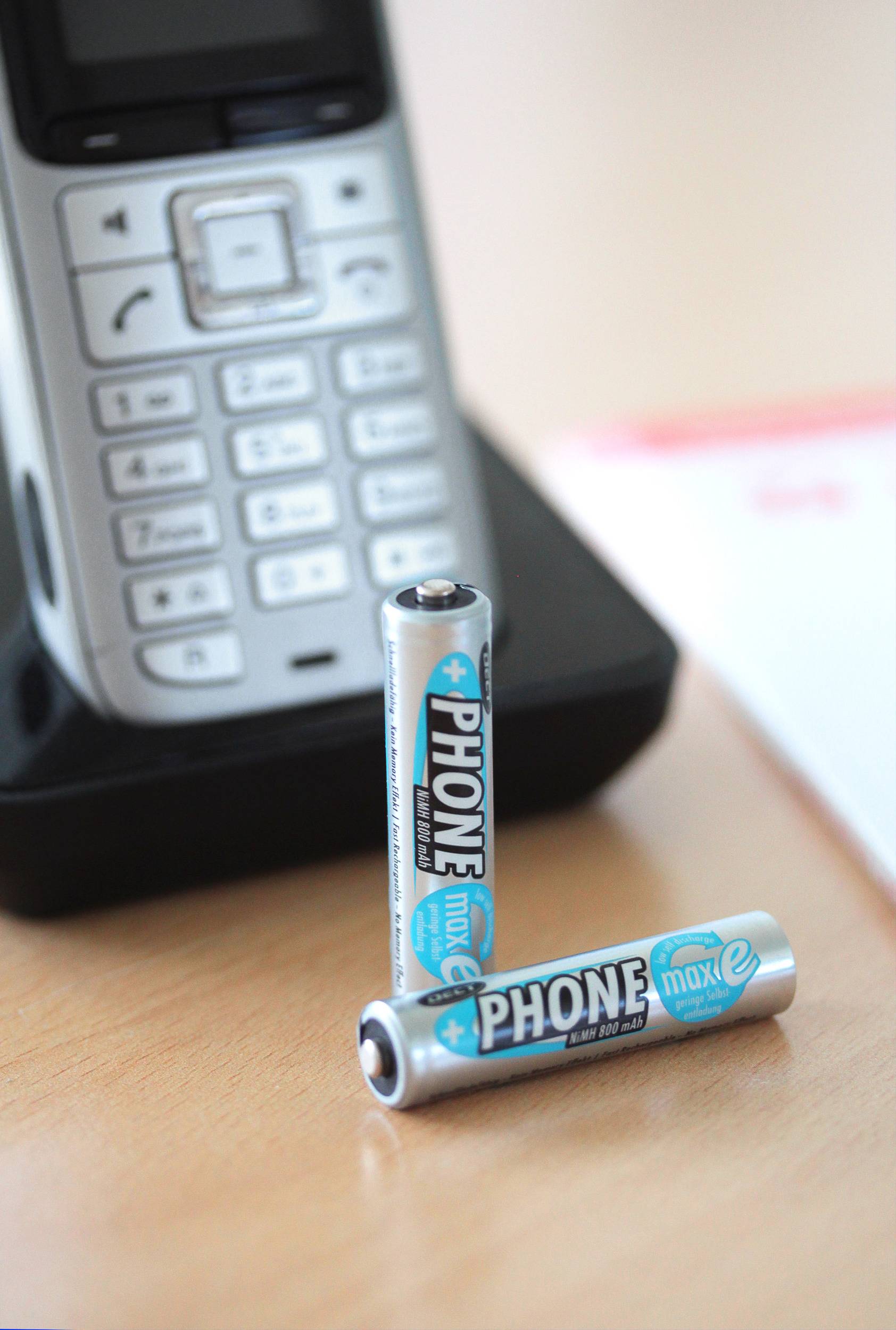 Před bezdrátovým telefonem leží dvě baterie s nápisem 'PHONE max e'.
