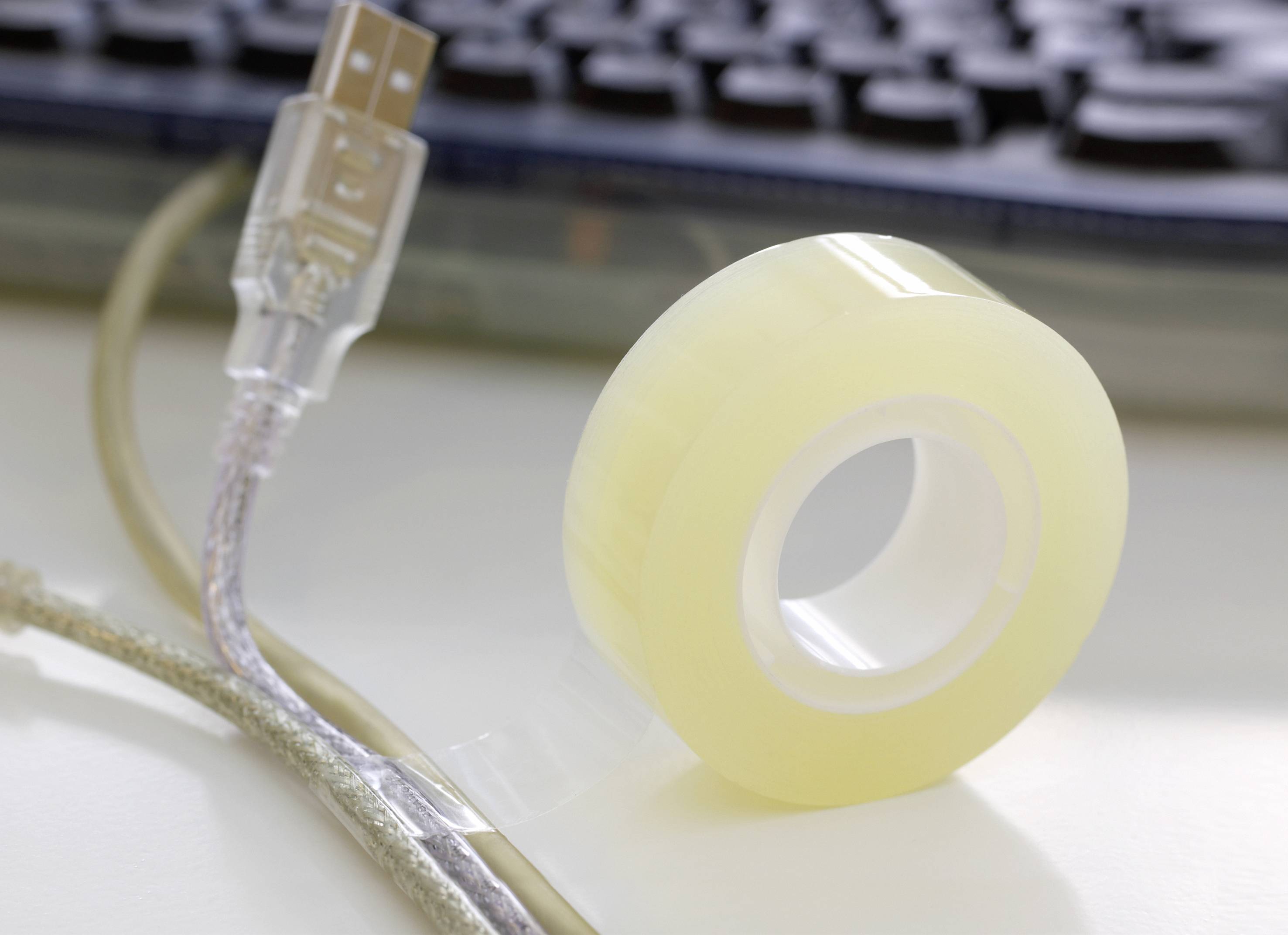 Role lepicí pásky a USB kabel na stole, v pozadí částečně zakryté počítačové klávesnice.