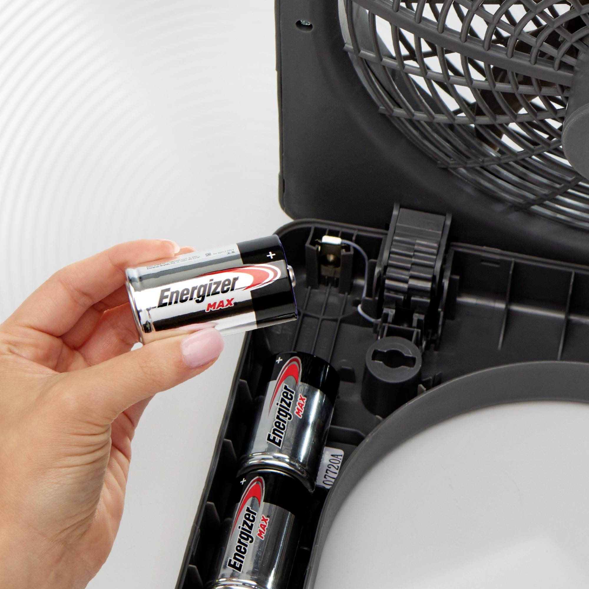 Osoba vkládá baterii AA značky 'Energizer Max' do prostoru pro baterie. V pozadí jsou další baterie.