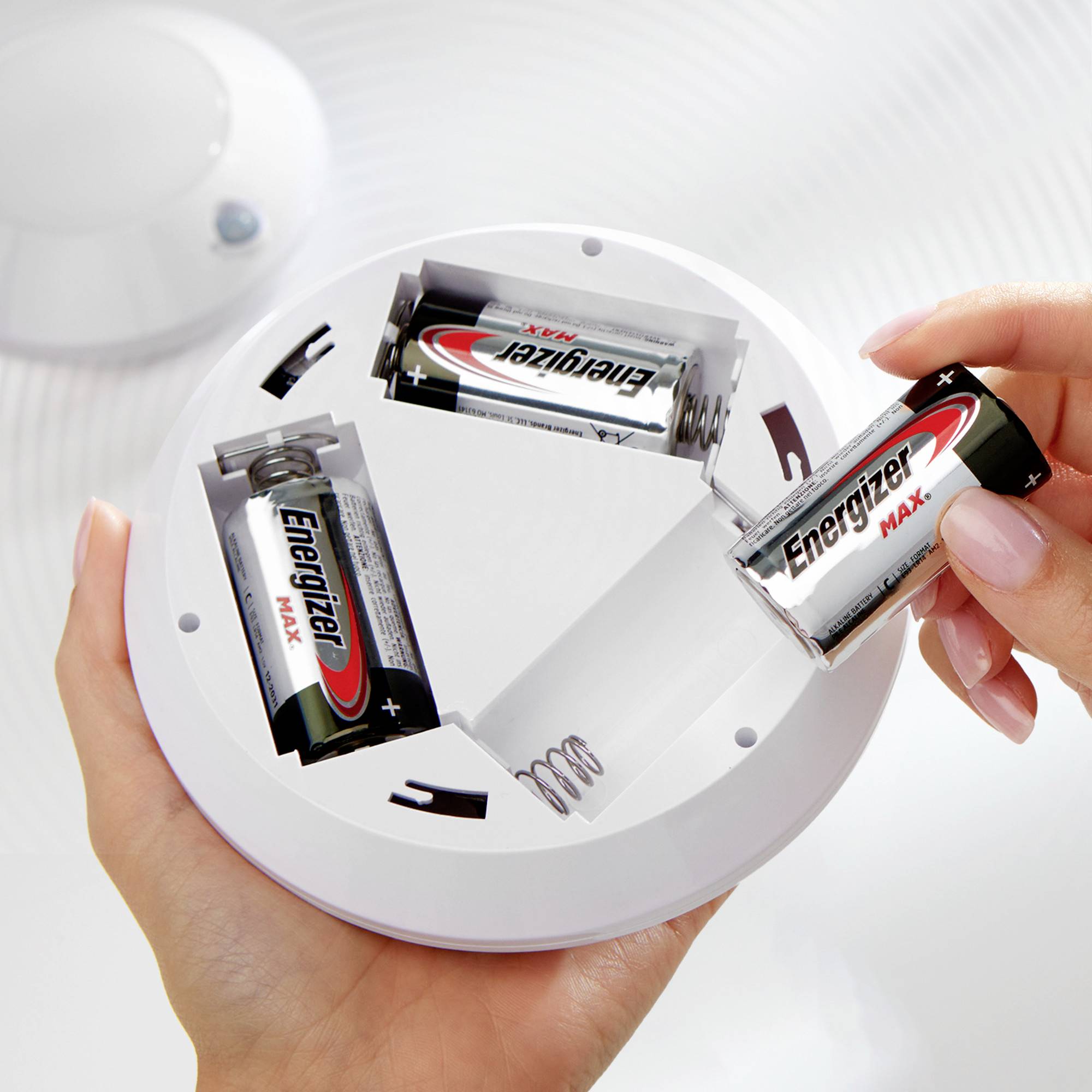 Dvě ruce drží detektor kouře, do kterého jsou vkládány baterie. Baterie jsou částečně vyjmuty a nesou logo společnosti Energizer.