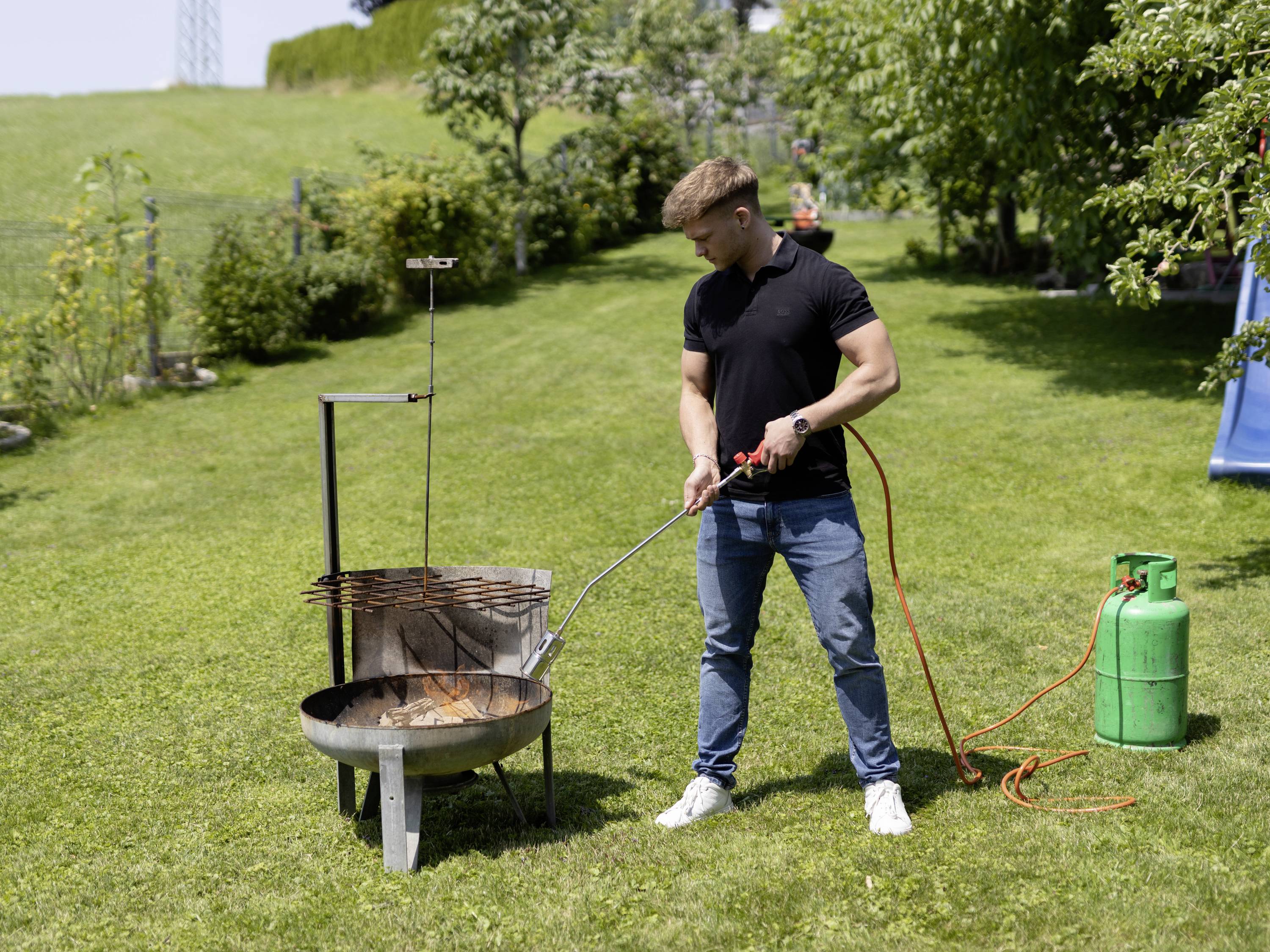 Muž v černém tričku a modrých džínách zapaluje plynový hořák na grilu v zelené zahradě.