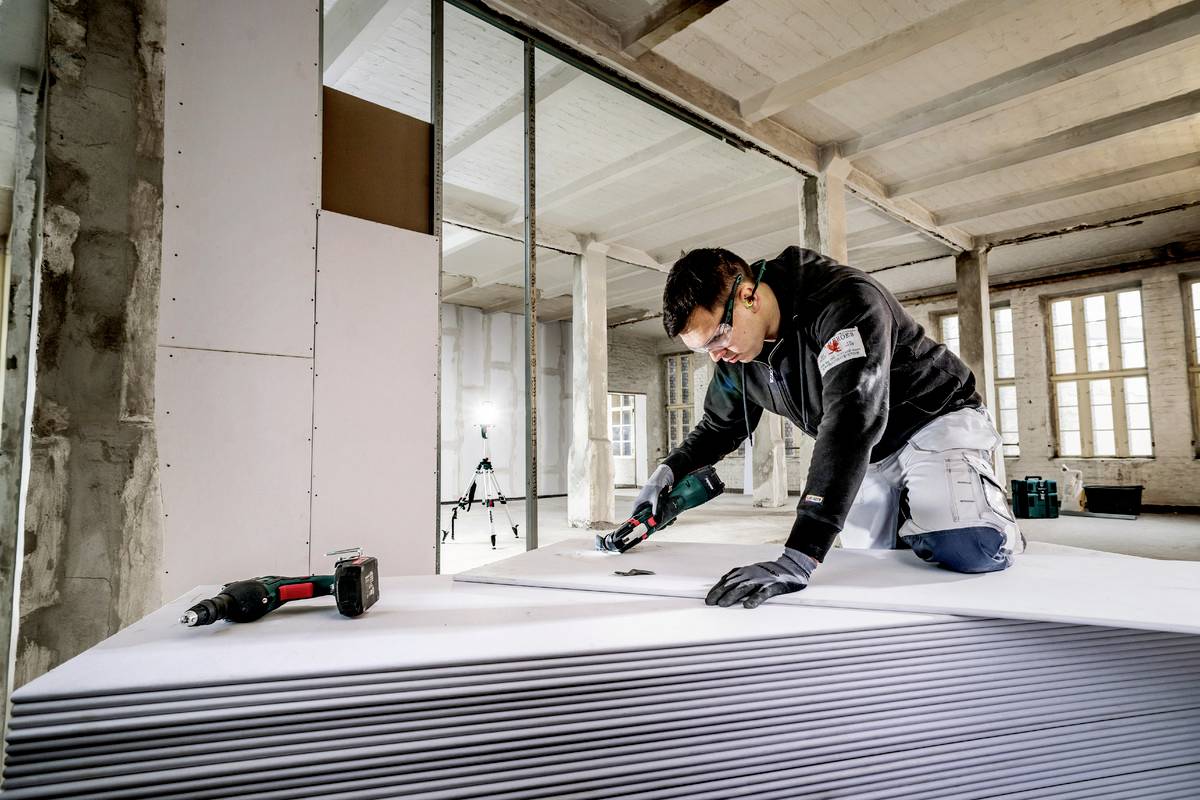 Řemeslník řeže sádrokartonové desky v projektu výstavby budovy. Má nasazené ochranné brýle a soustředěně pracuje s elektrickým nástrojem.