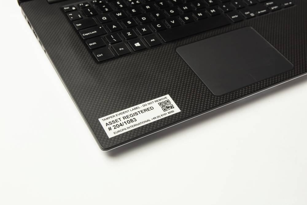 Notebook s černou klávesnicí zobrazuje štítek s nápisem 'Asset Registered' a sériovým číslem na opěrce zápěstí.