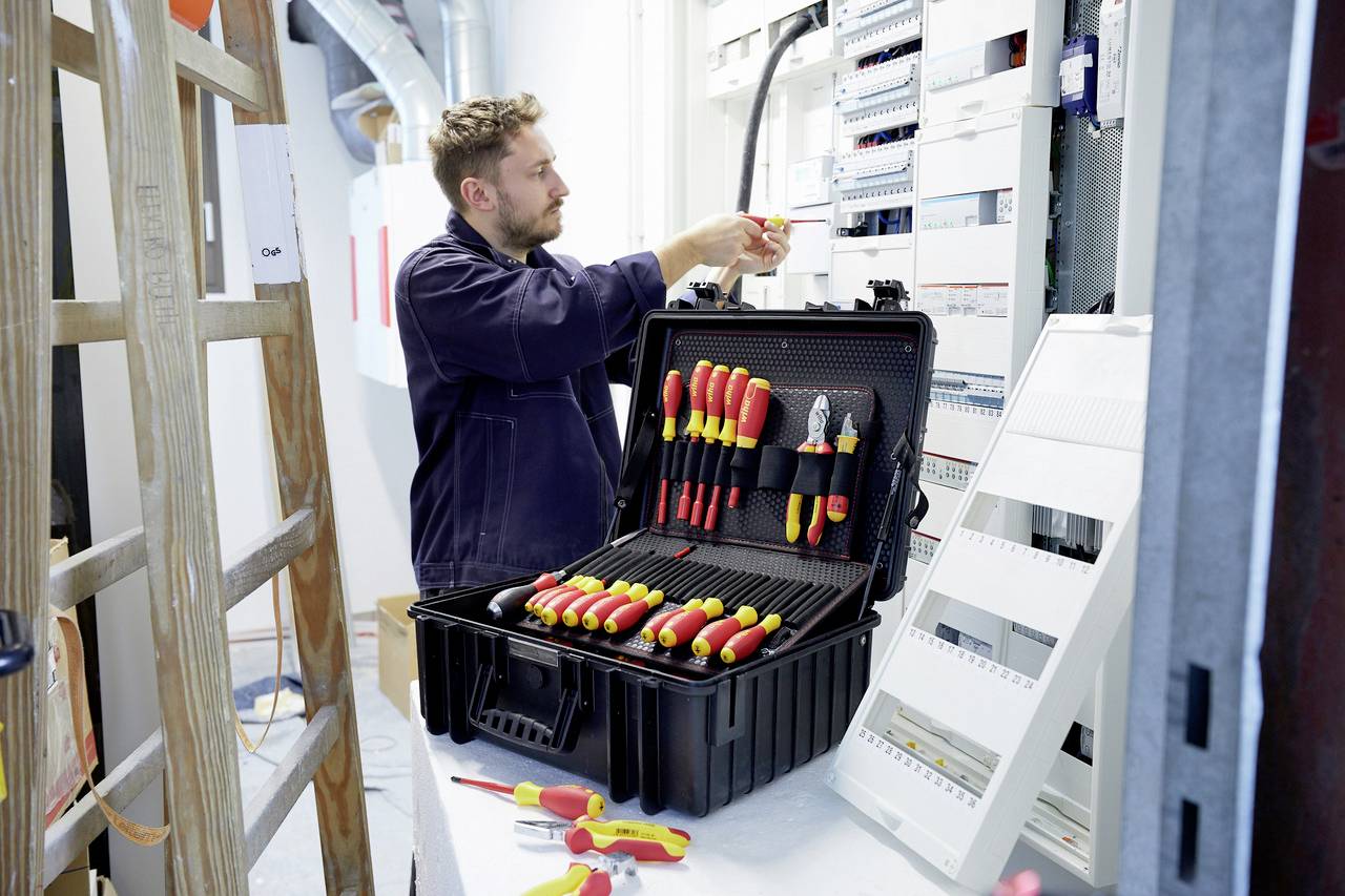 Elektrikář pracuje na rozvodné skříni s otevřenou brašnou plnou šroubováků a kleští. Žebřík stojí vedle něj.