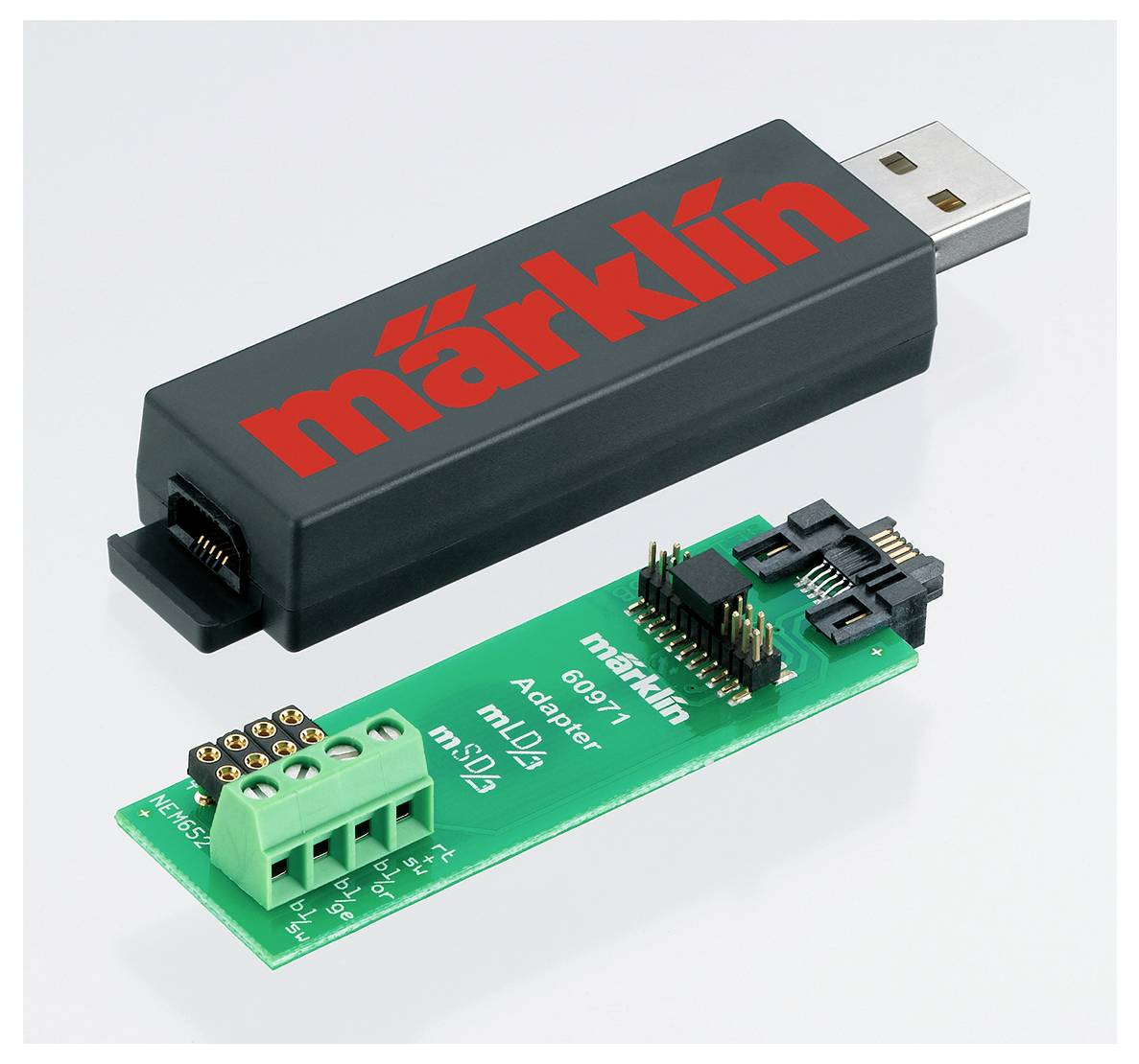 USB zařízení s logem 'märklin' a zelená adaptérová deska s několika konektory, obojí pro řízení modelové železnice.