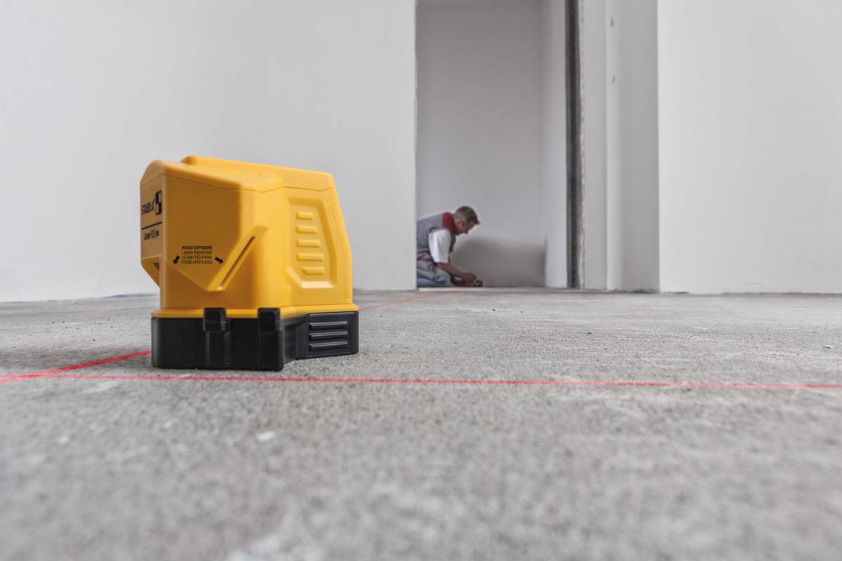 Žluté laserové nivelační zařízení promítá čáry na betonovou podlahu v prázdné místnosti; muž pracuje v pozadí.