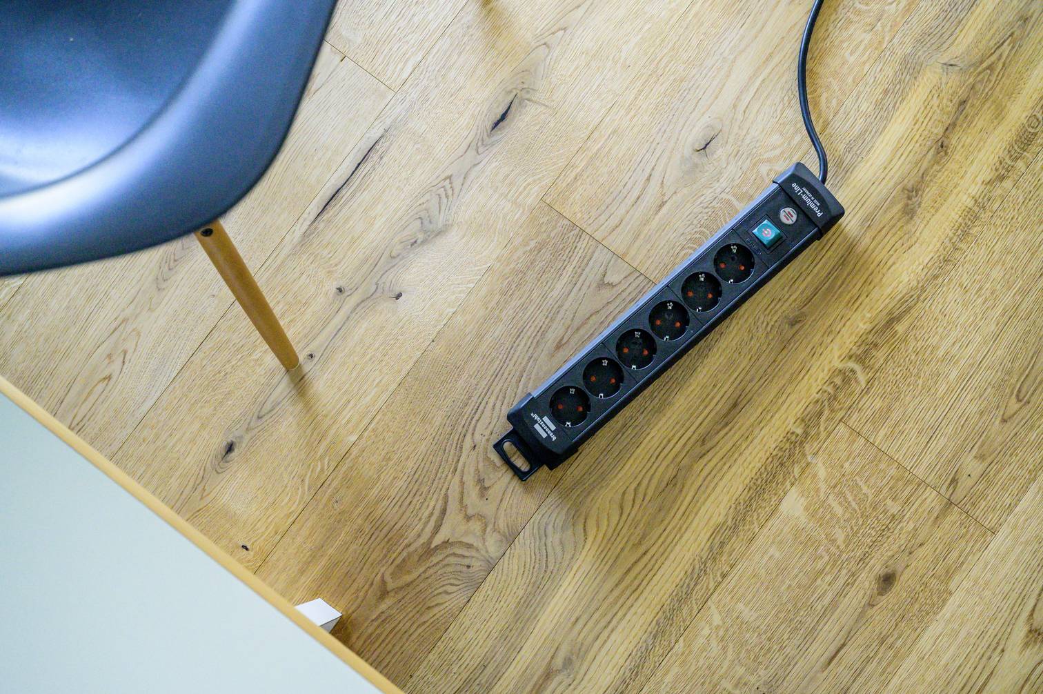 Černá prodlužovací zásuvka se šesti místy leží na dřevěné podlaze vedle nohy stolu.