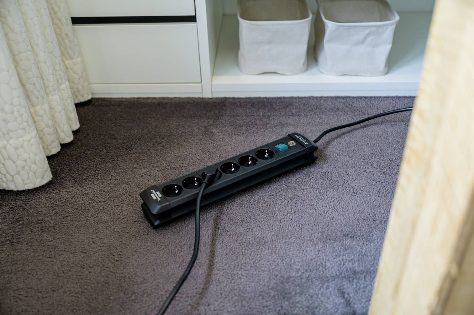 Černá prodlužovací lišta leží na koberci před otevřenou policí se dvěma textilními košíky. Kabel vede mimo záběr.