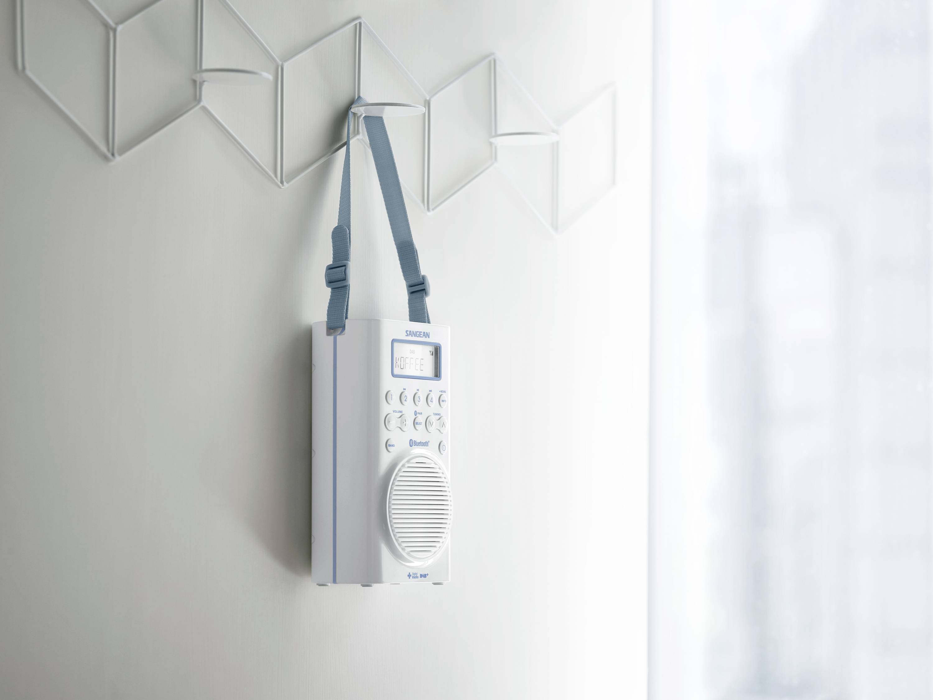Přenosné modro-bílé rádio visí na šedé zdi. Vedle něj je geometrická nástěnná dekorace a rozmazané pozadí.