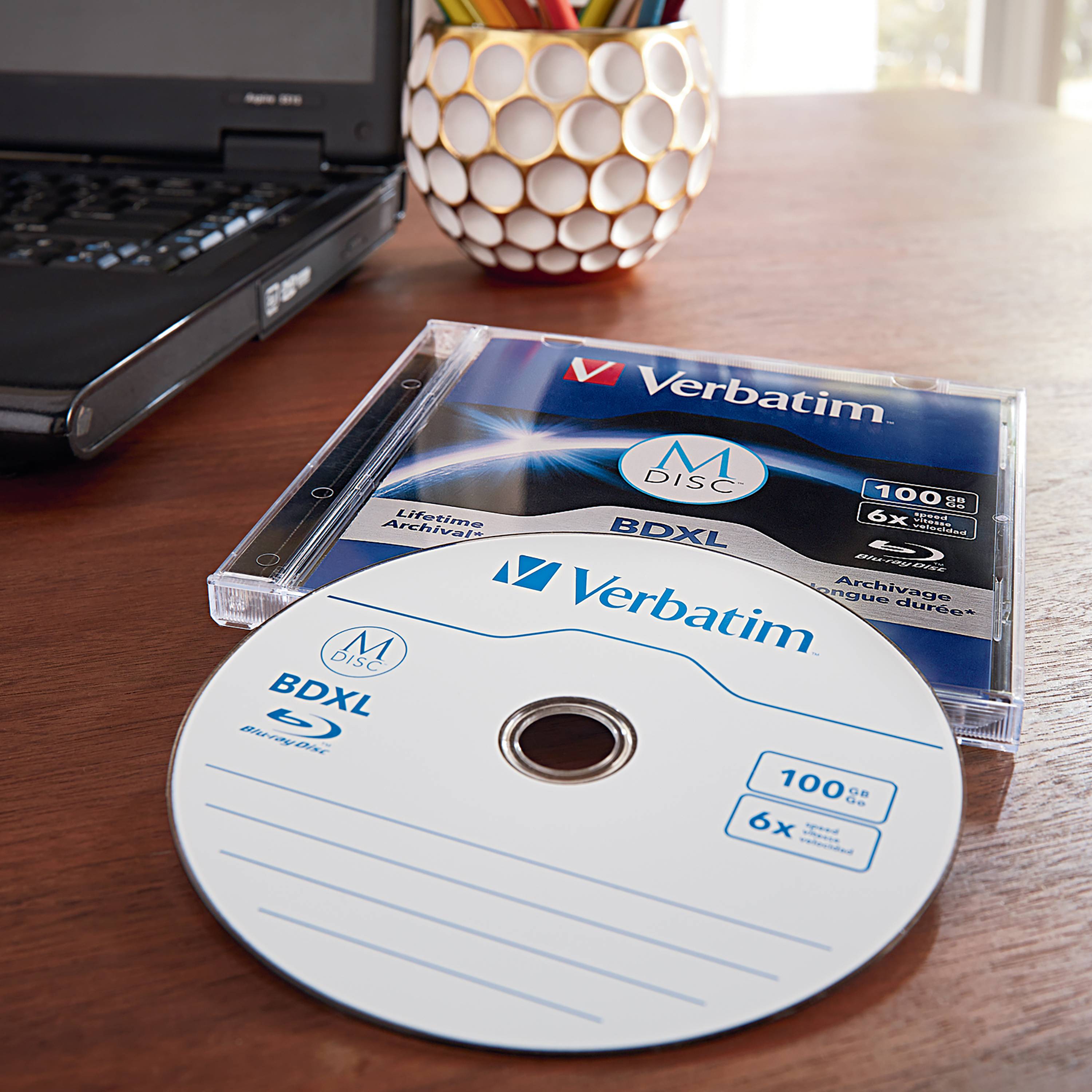 Laptop leží na stole vedle Verbatim Blu-ray disku v obalu. V pozadí se nachází stojánek na tužky.