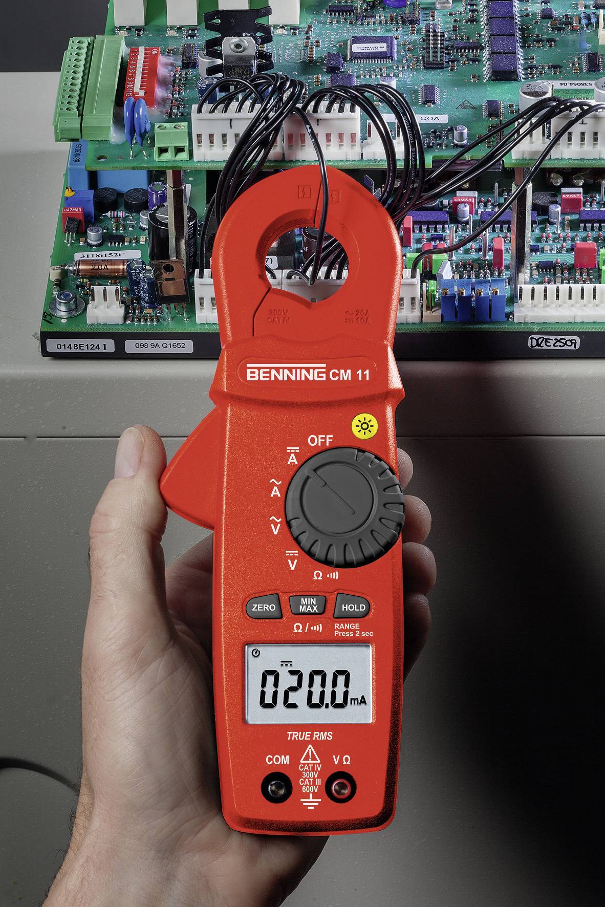 Ruka drží červený multimetr připojený k desce plošných spojů, který ukazuje 20,0 mA. V pozadí jsou viditelné elektronické komponenty.
