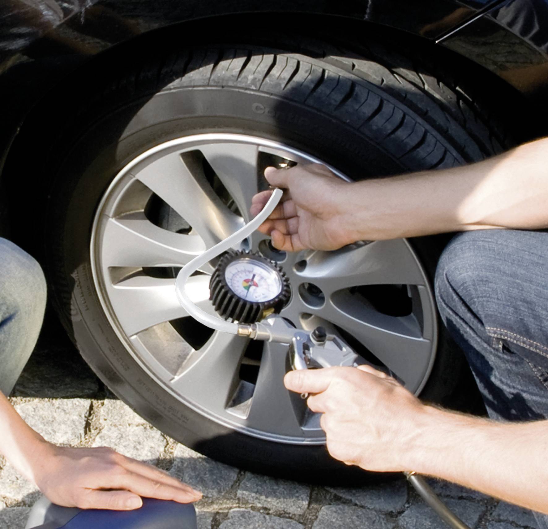 Dva lidé kontrolují a huští pneumatiku automobilu pomocí tlakoměru. Pneumatika je umístěna na dlážděné ploše.