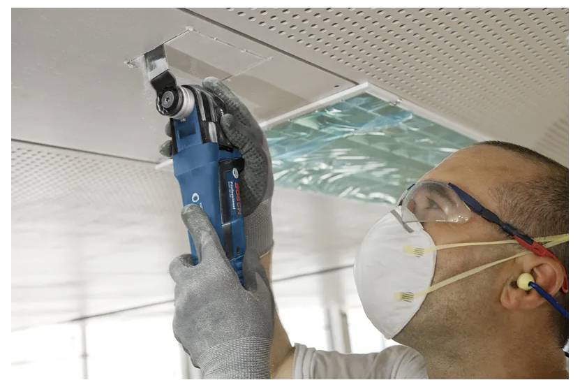 Technik v rukavicích, roušce a ochranných brýlích kontroluje větrací otvor v stropě nebo panel pomocí modrého ručního zařízení.