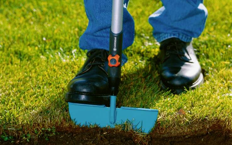 Osoba v modrých džínách a černých botách používá ruční modrý ořezávač trávníku k zastřihávání trávy podél okraje trávníku.