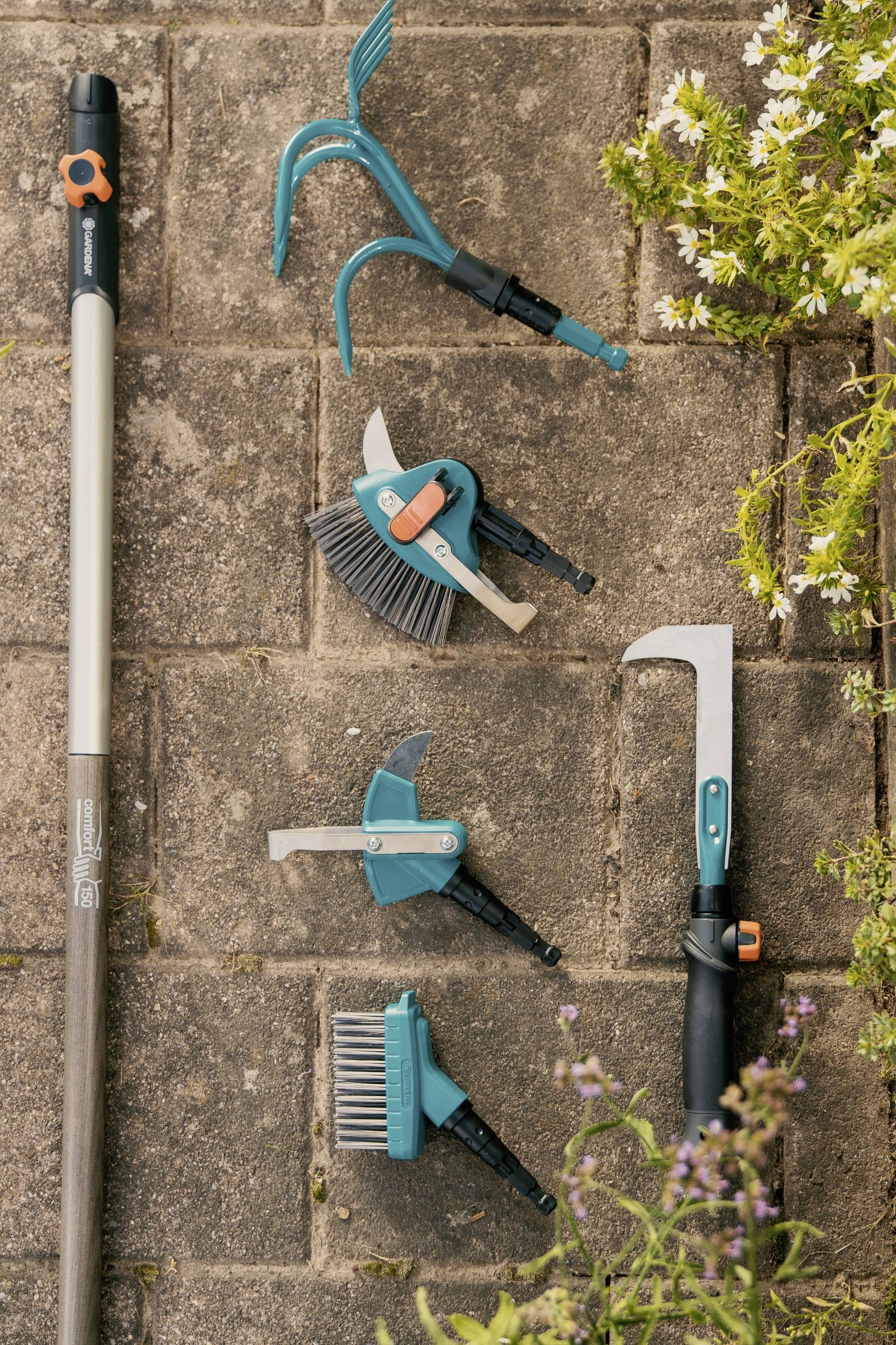 Zahradní nářadí na kamenné podlaze: rukojeť lopaty, hrábě, kartáč, spárový čistič, vytrhávač plevele, obklopené kvetoucími rostlinami.