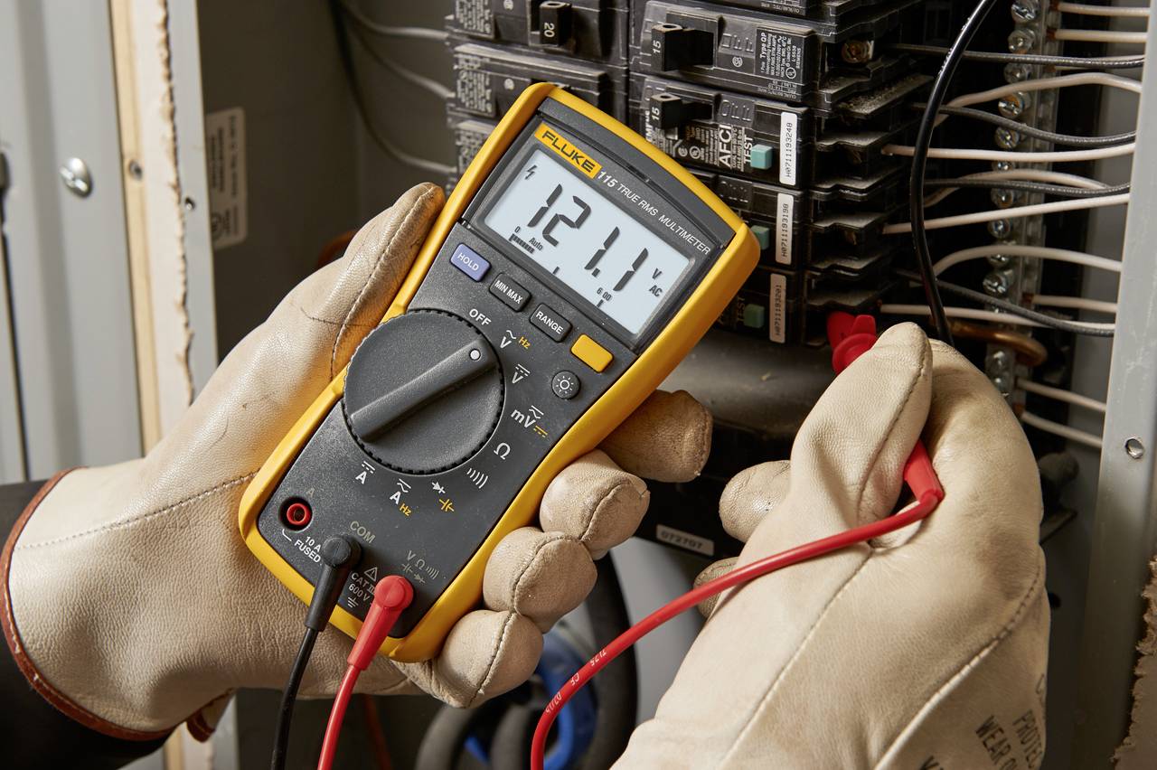 Jedna osoba měří digitálním multimetrem napětí 121,1 voltů na elektrickém rozvaděči.