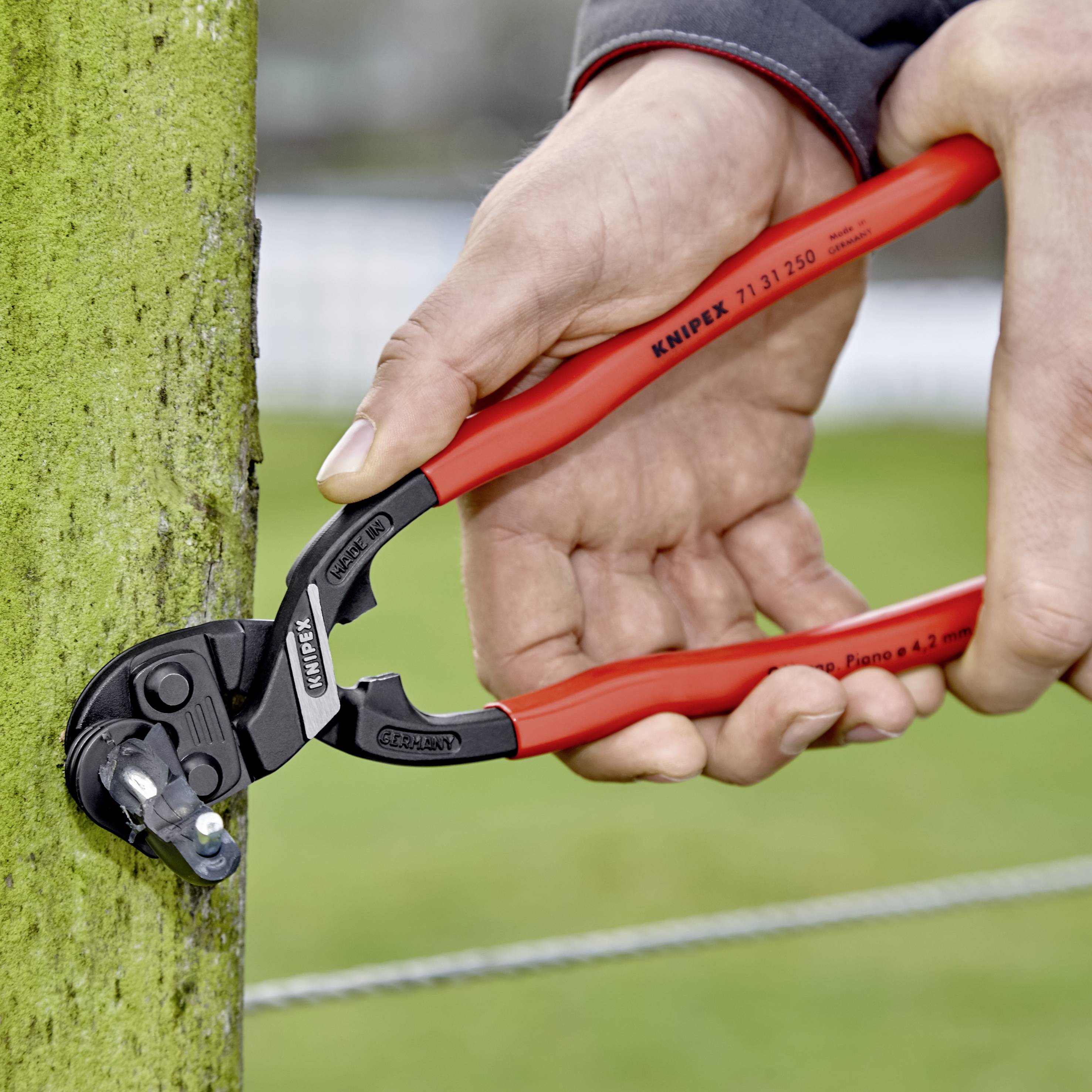 Osoba používá červené kabelové štípací kleště Knipex k odstřižení zeleného kabelu ve venkovním prostoru.