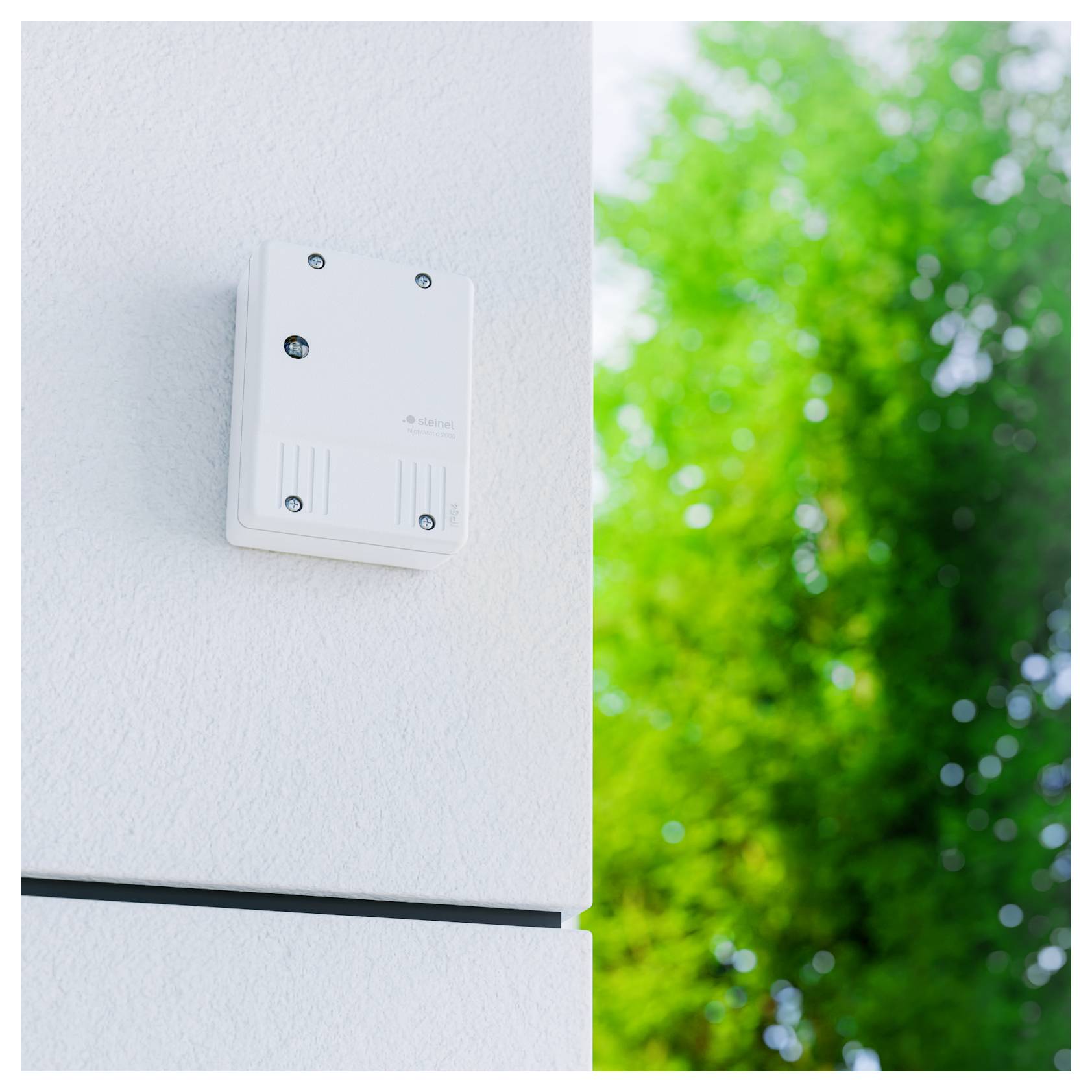 Malý bílý nástěnný snímač nebo ovládací box umístěný na vnější stěně s rozostřenými zelenými stromy v pozadí.