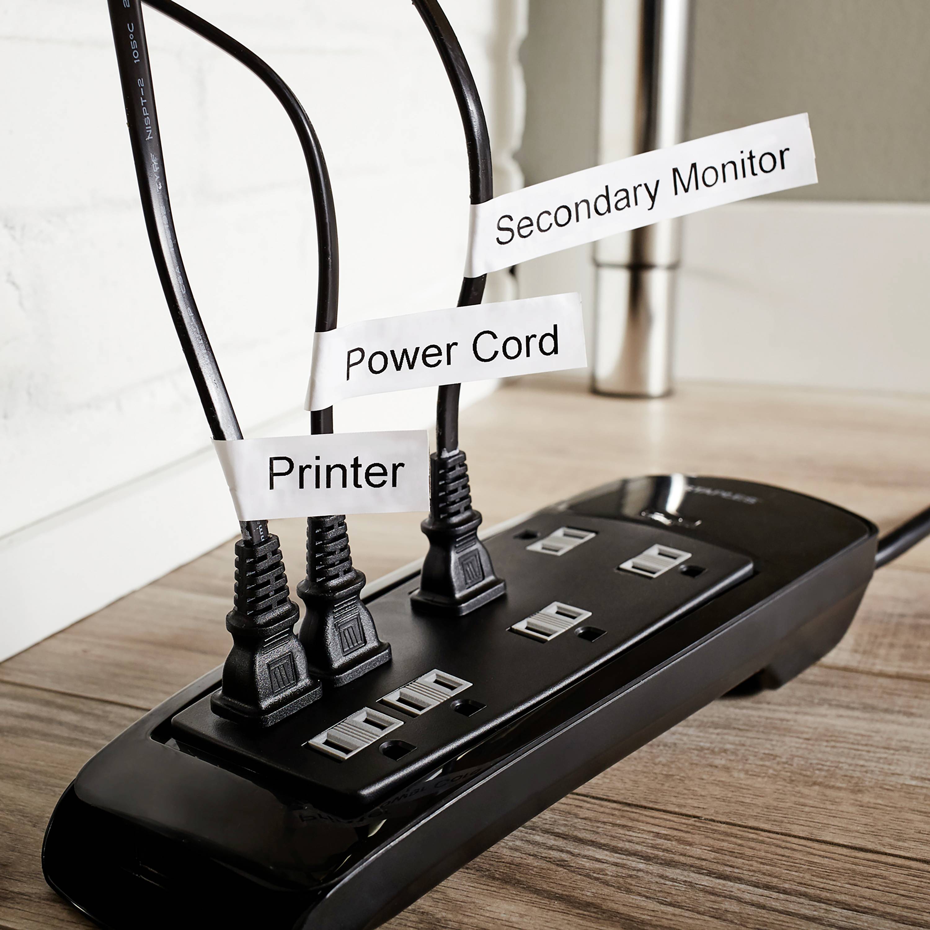 Prodlužovací lišta na dřevěné podlaze se třemi zapojenými kabely; označená štítky 'Sekundární monitor', 'Napájecí kabel', 'Tiskárna'.