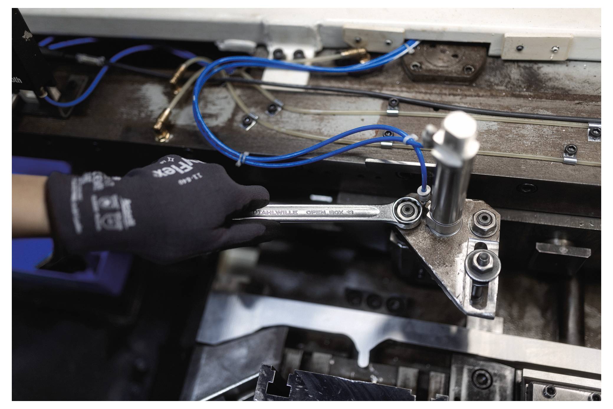 Osoba v rukavici používá klíč na průmyslovém stroji s viditelnými modrými vodiči, což naznačuje údržbu nebo mechanické práce.