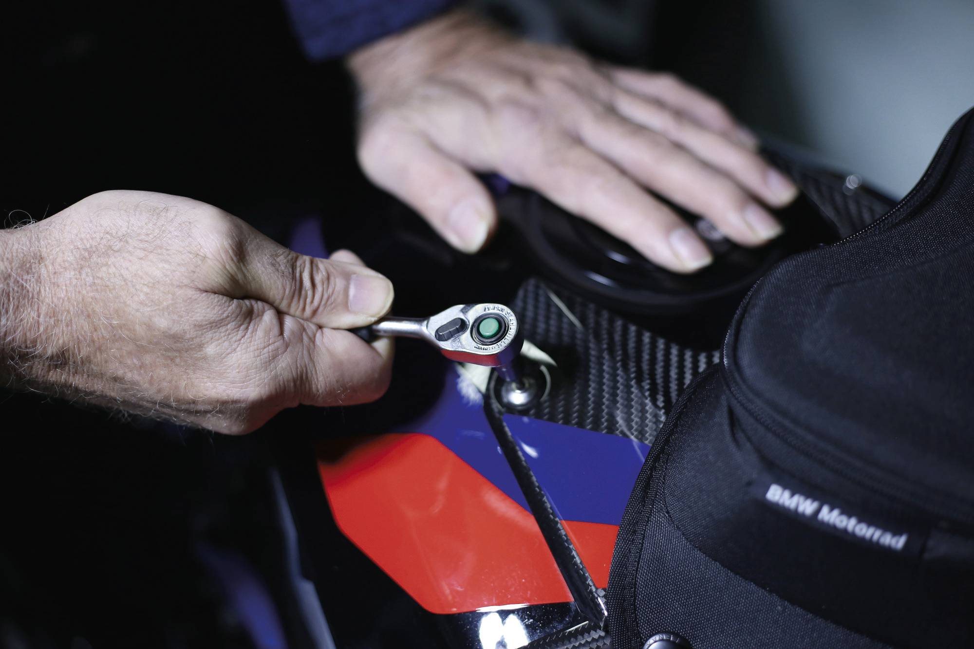 Osoba používá klíč k seřízení části motocyklu, přičemž předvádí údržbu vozidla se zaměřením na nástroje a ruce.