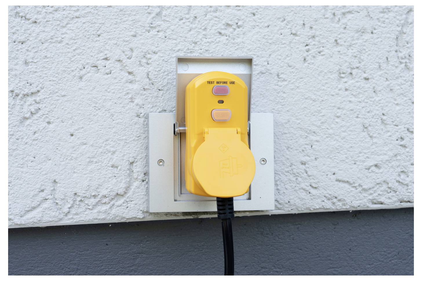 Žlutý kryt venkovní elektrické zásuvky připevněný na stěně, se síťovým kabelem zasunutým a visícím dolů.