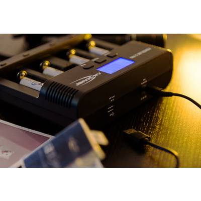 Nabíječka se čtyřmi vloženými bateriemi AA a modrým displejem, zobrazující stav nabití, obklopená USB kabelem a rozmazaným pozadím.