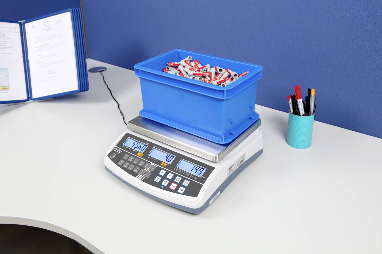 Malá modrá krabice s trubkami na elektronické váze. Displej ukazuje 5,460 kg, 40 jednotek, 149 eur. Kancelářské pozadí.