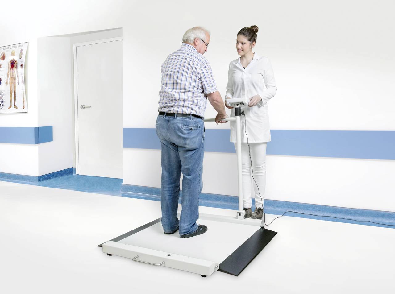 Muž je doprovázen ženou na váhu v ordinaci lékaře. Na stěně visí anatomický plakát.