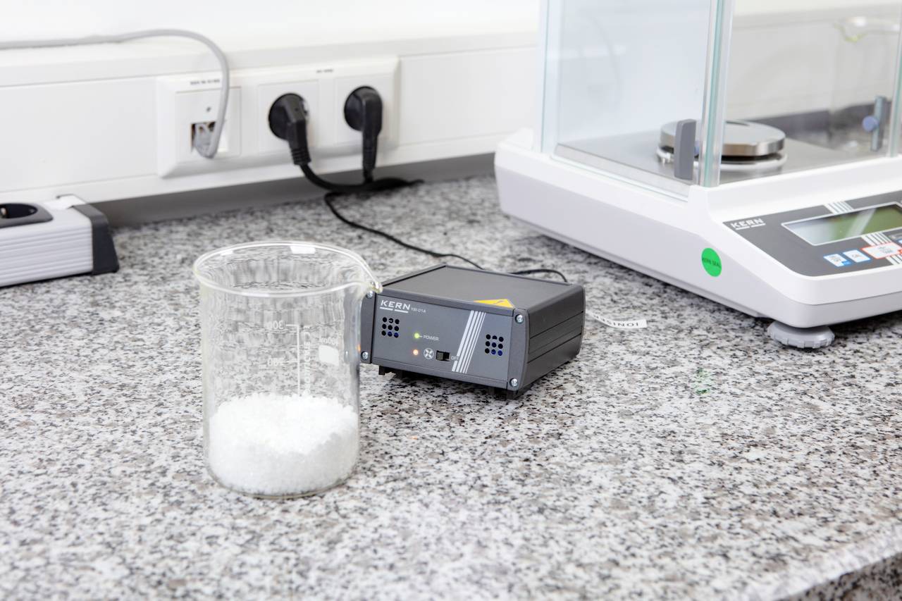 Nádoba s bílým práškem stojí na laboratorním stole vedle elektronického přístroje a váhy v laboratorním prostředí.