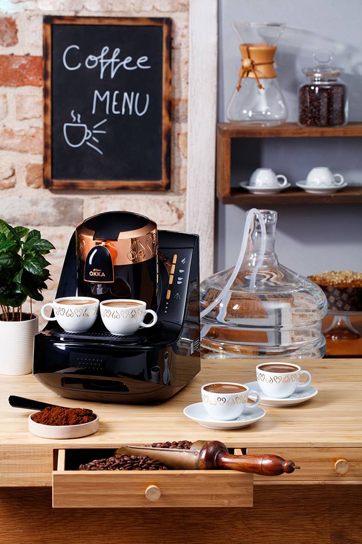 Kávový přístroj na dřevěné polici, obklopený hrnky, džbánem na vodu a kávovými zrny. V pozadí cedule s nápisem 'Coffee Menu'.