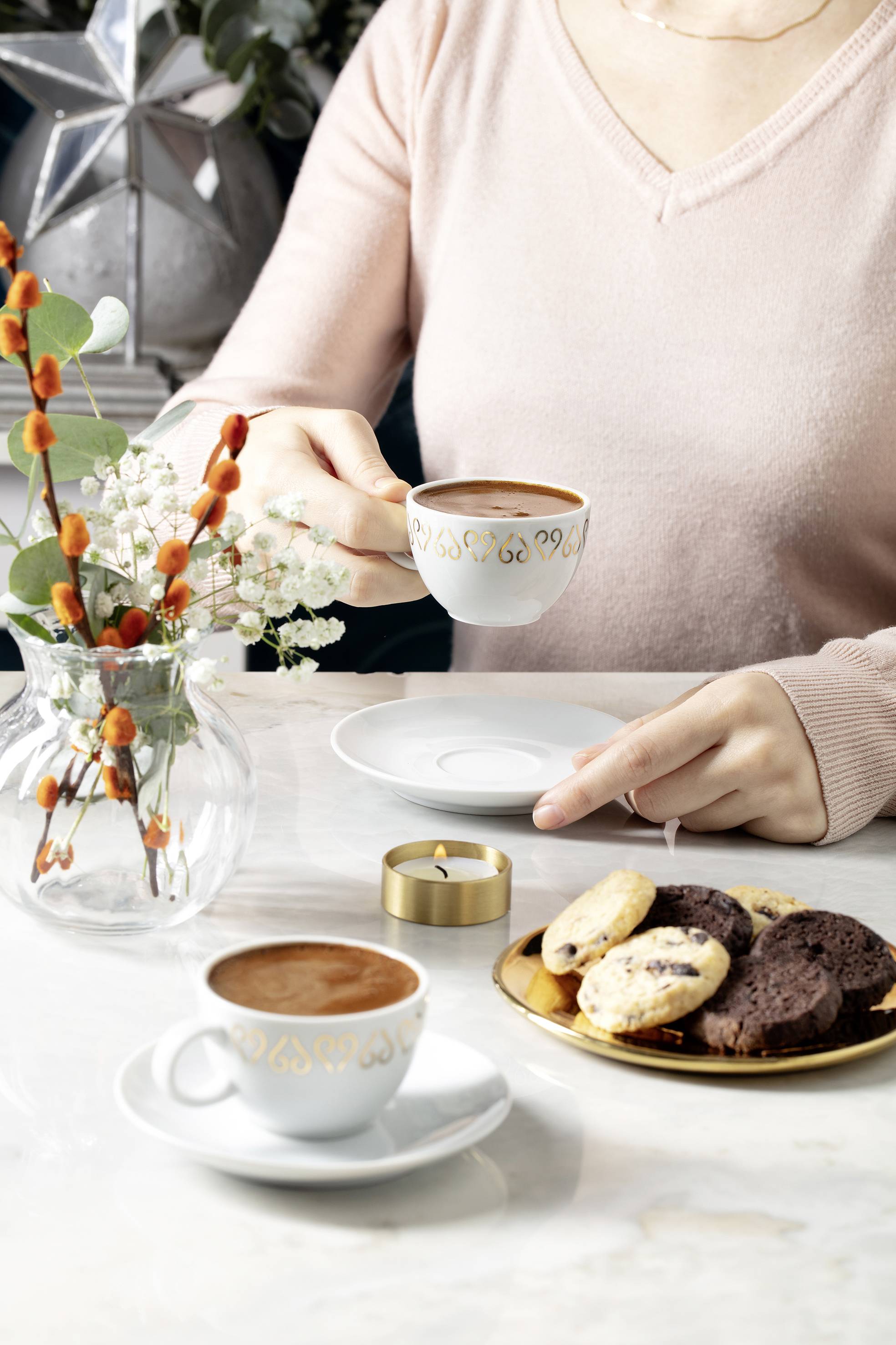 Osoba drží šálek kávy, vedle stojí další káva a talíř s cookies na stole. V pozadí je váza s květinami.