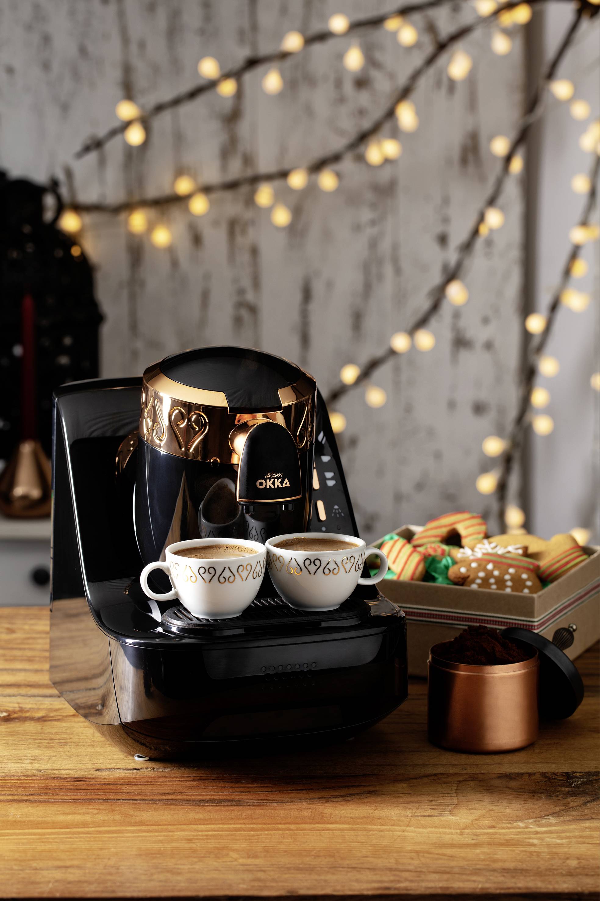 Na stolku stojí černý kávovar připravující dvě espresso šálky. V pozadí jsou vidět světelné řetězy a barevné pečivo.