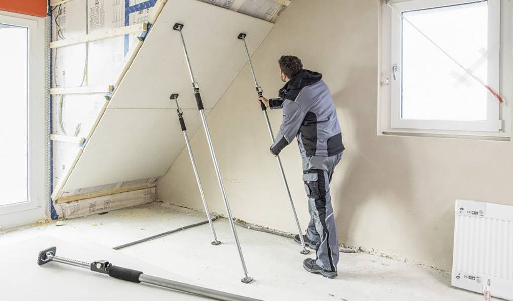 Řemeslník upevňuje sádrokartonovou desku pomocí vzpěr na šikmé střeše. V místnosti je viditelné okno a topné těleso.