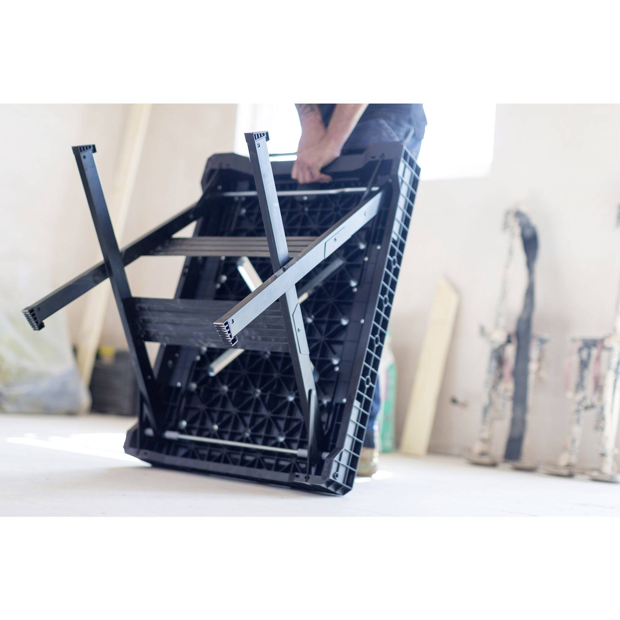 Osoba montuje nohy převráceného stolu v jasně osvětlené místnosti. V pozadí jsou vidět malířské potřeby.