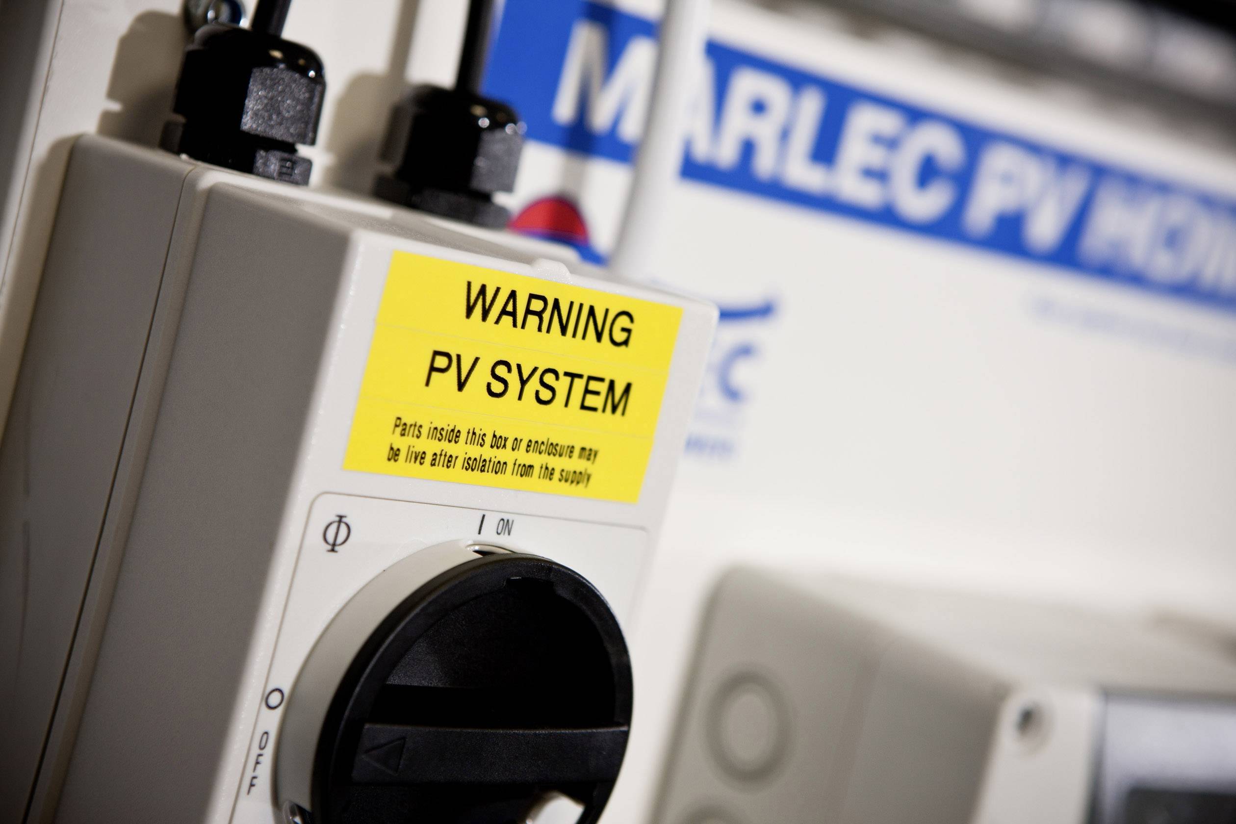 Výstražná cedule vedle komponenty fotovoltaického systému s varováním 'WARNING PV SYSTEM'. V pozadí rozmazaná samolepka Marlec PV. Hlavní zaměření na bezpečnostní upozornění.