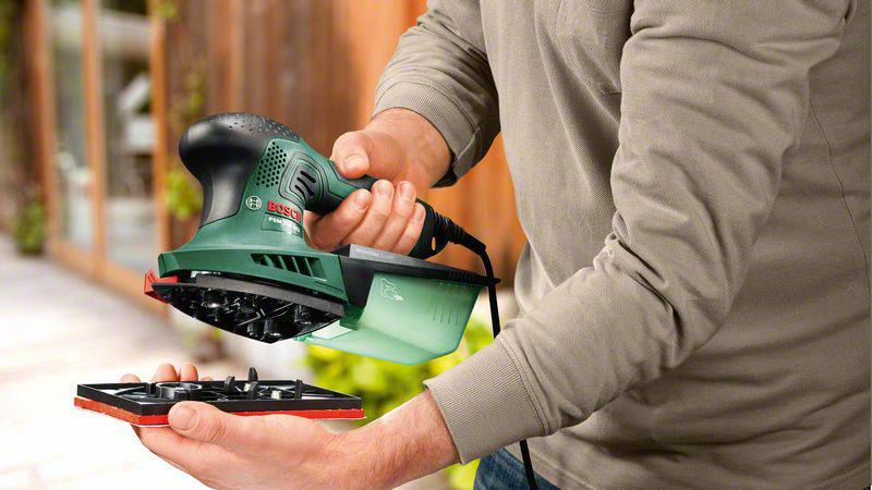 Osoba drží zelený elektrický nástroj, který se podobá brusce, před rozmazaným pozadím dřevěného domu.