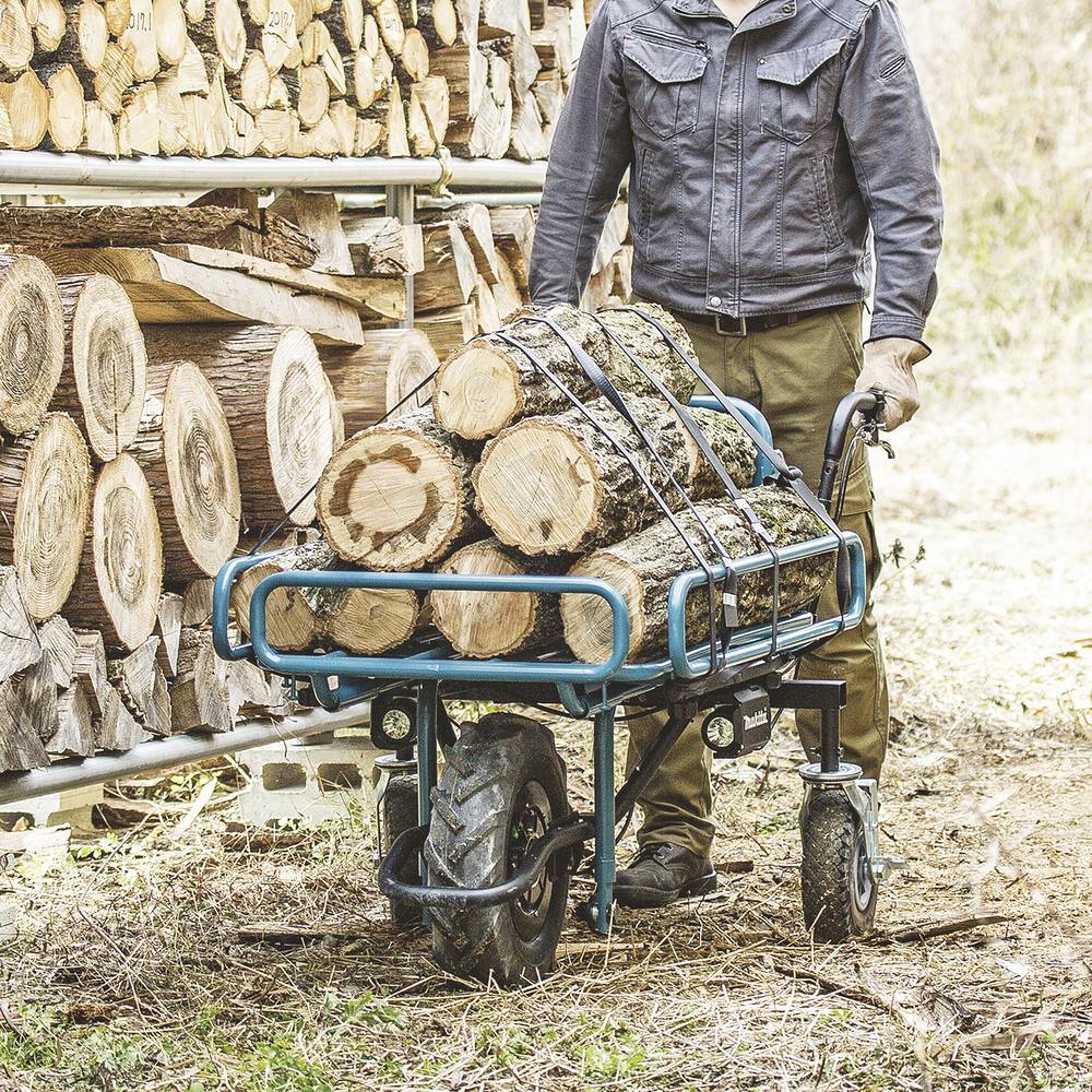 Osoba v pracovním oblečení tlačí dřevěný vozík plný nařezaných kmenů vedle hromady dřeva venku.