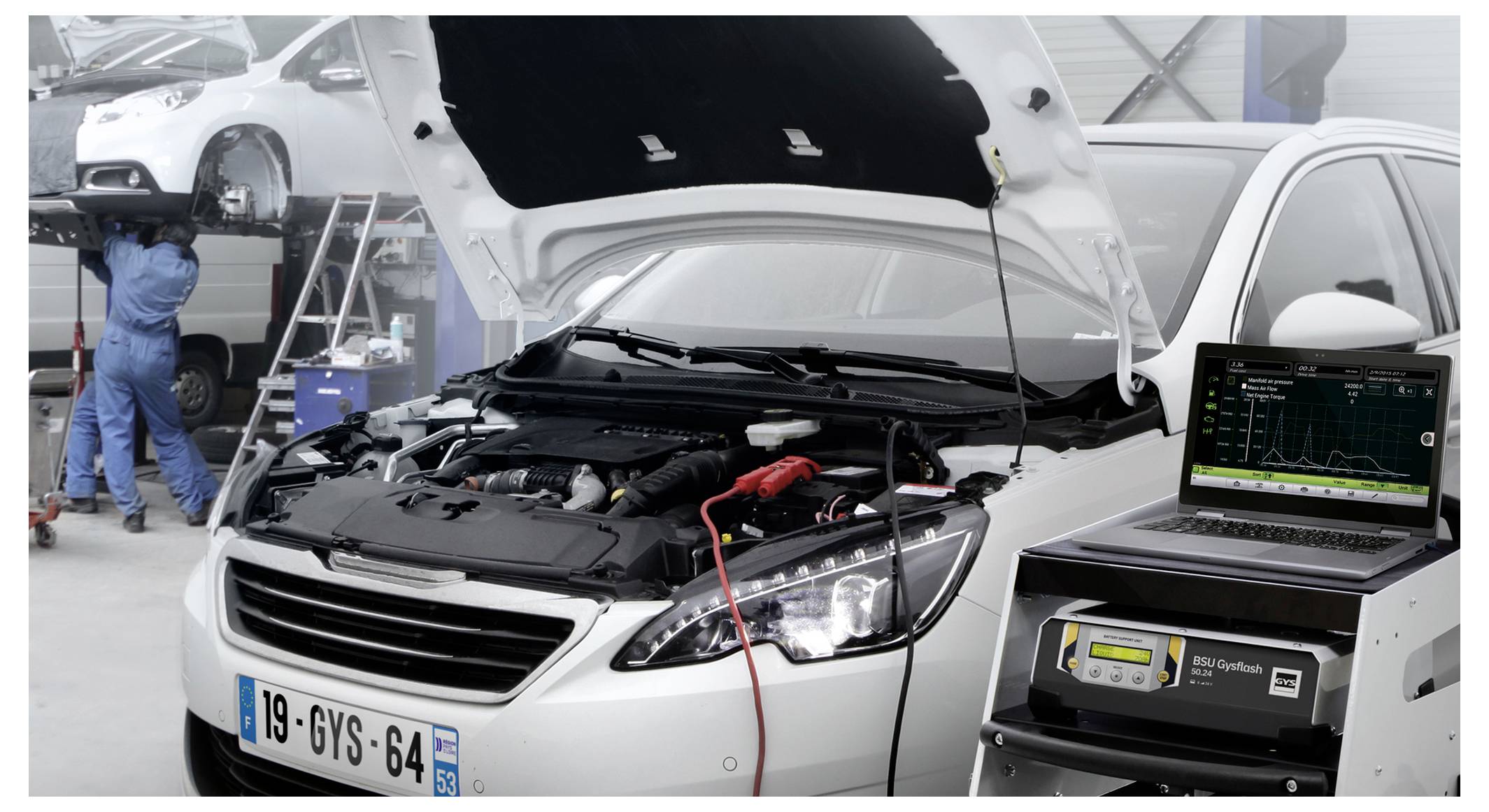 Auto v autoopravně s otevřenou kapotou, baterie se testuje z diagnostické jednotky připojené k notebooku, zatímco v pozadí mechanic pracuje na jiném vozidle.